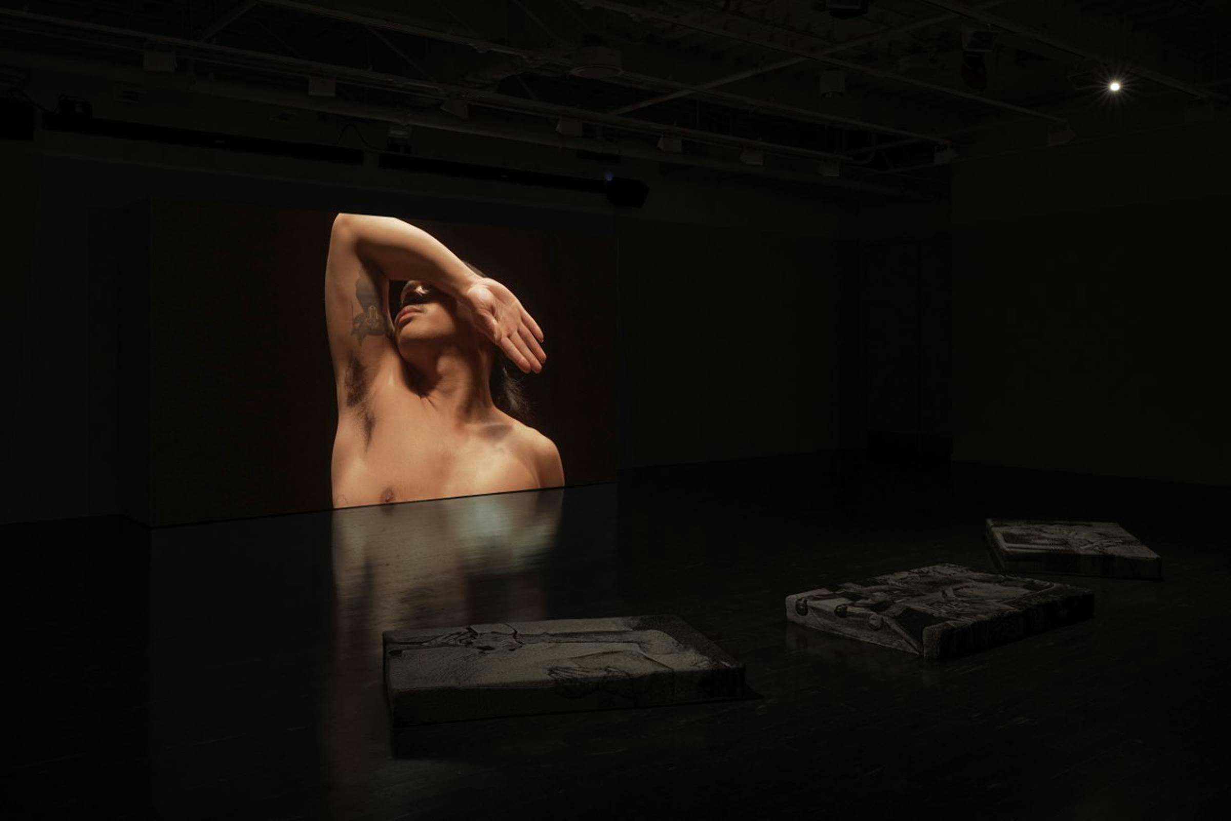 Installation view of The Heart of a hand in a darkened space. The image projected is of a shirtless person standing in a black space with their arm over their face.
