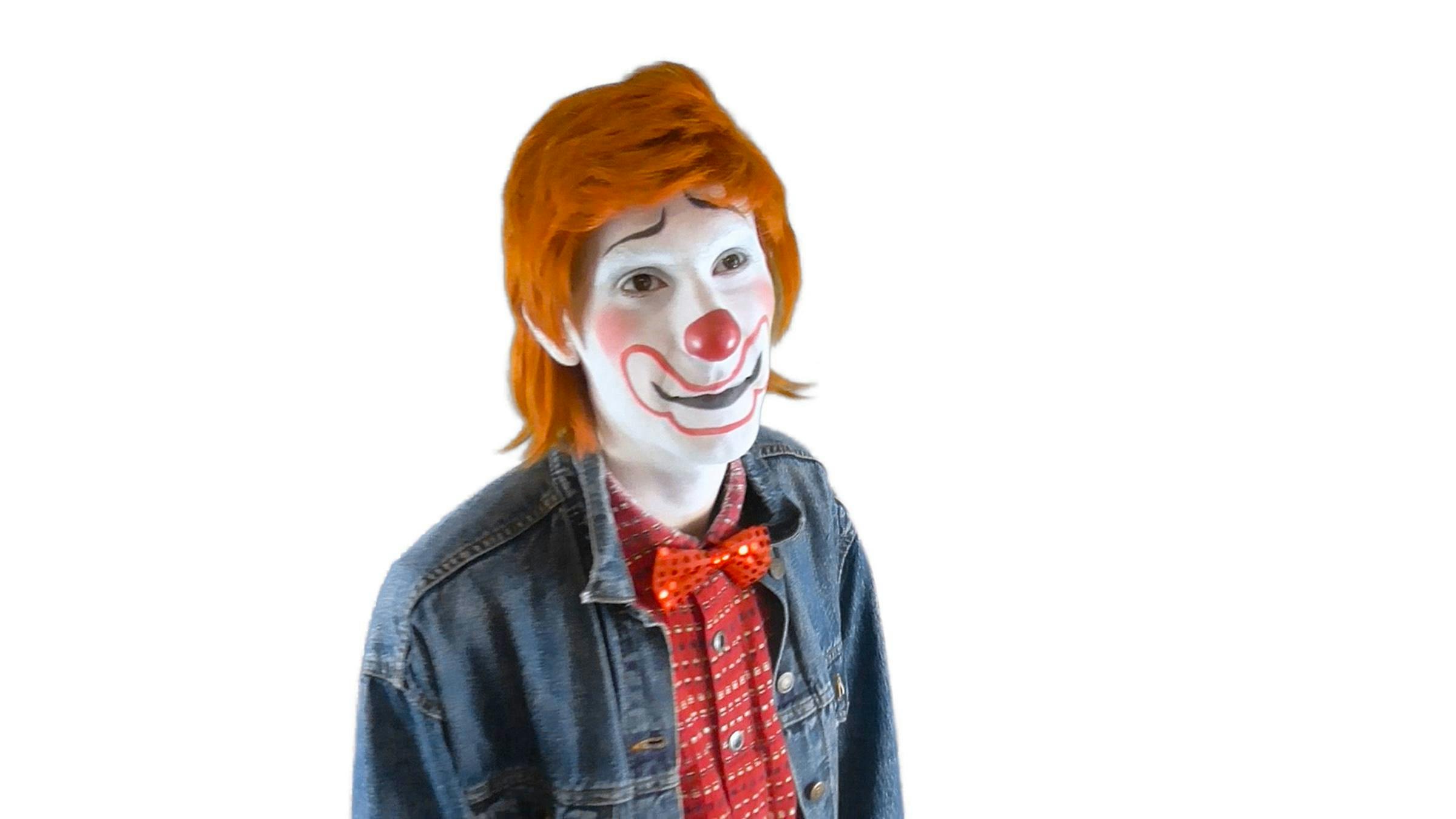 A person wearing clown-like make up, a red nose, orange hair and a red sparkling bow tie. The person is against a white background.