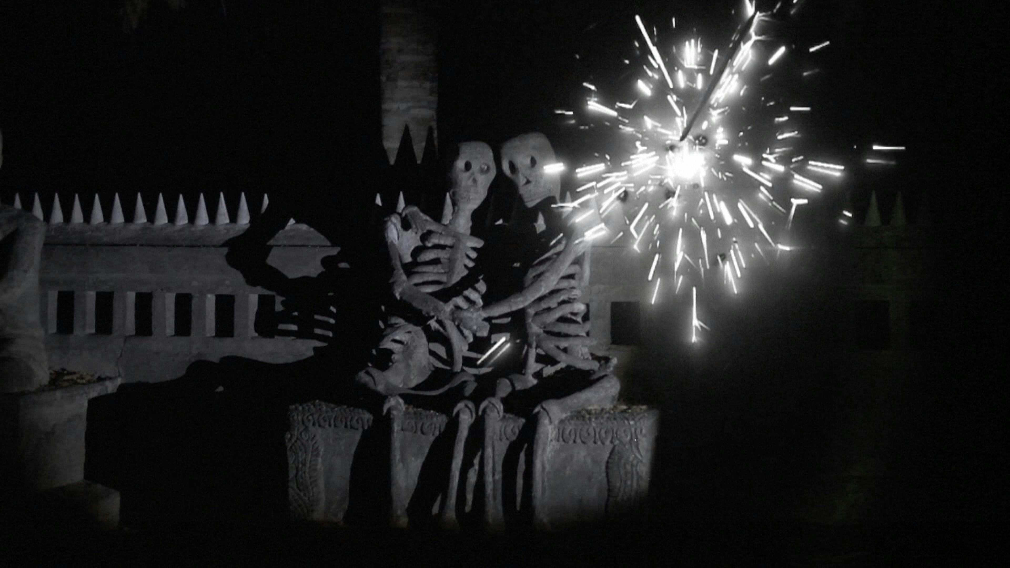 A black and white image of two skeletons are sitting on a stone bench embracing. There is a firework exploding next to them.
