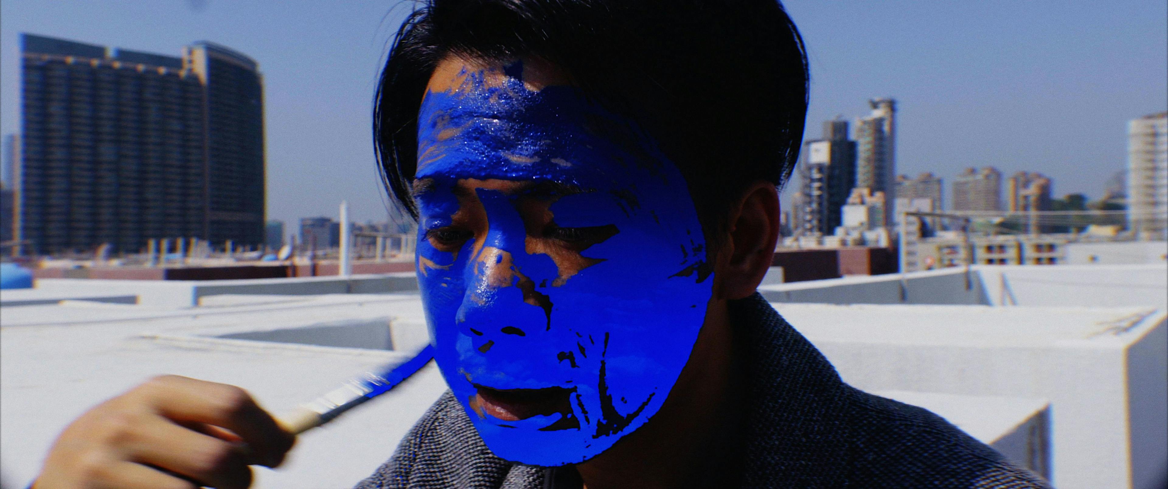 Close-up image of a person painting their face blue. In the background, there are many skyscrapers.
