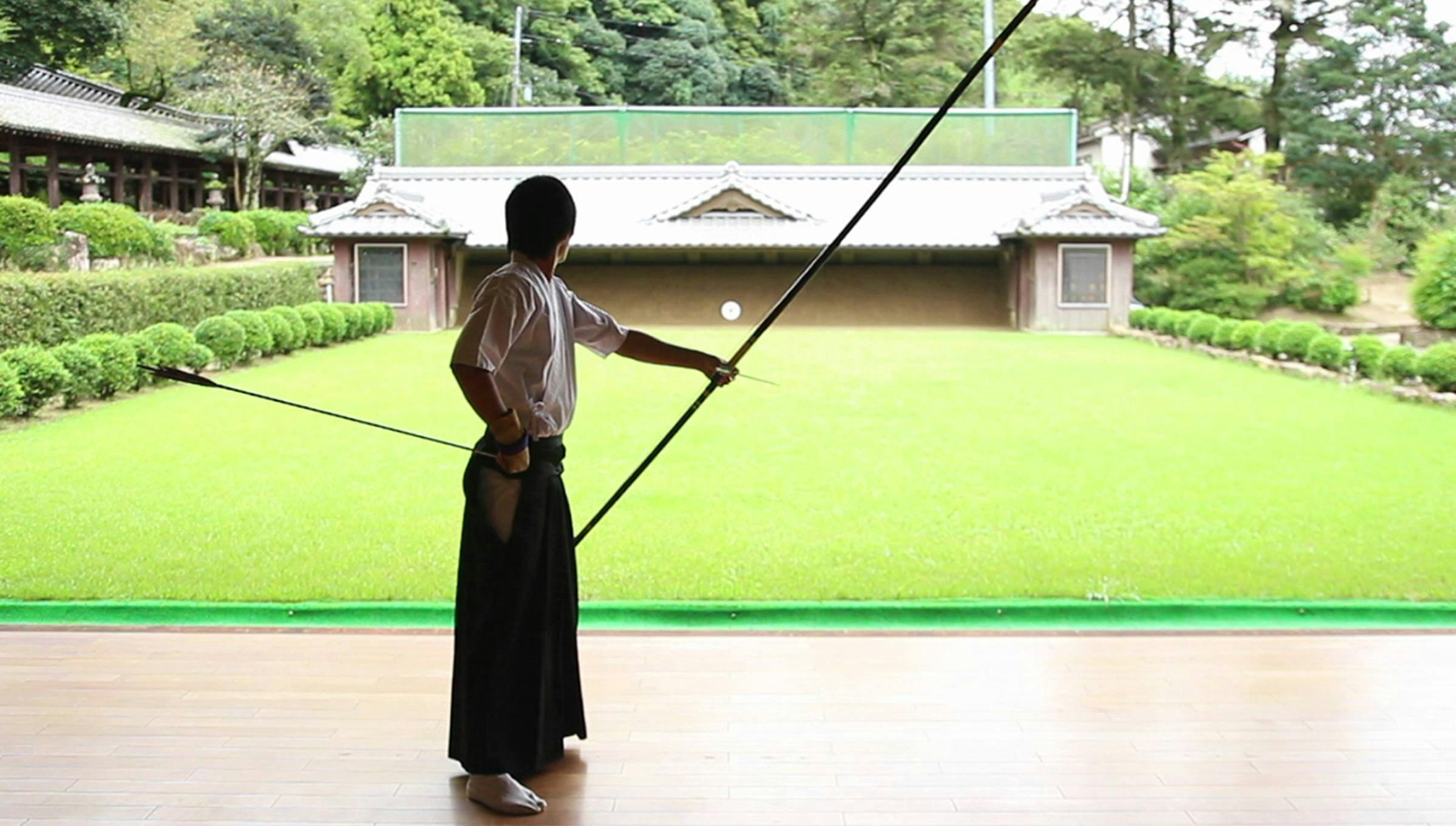 A person stands with their back to the camera holding a large bow in one hand and an arrow in the other. They stand in front of a grassy plane lined with formal bushes.