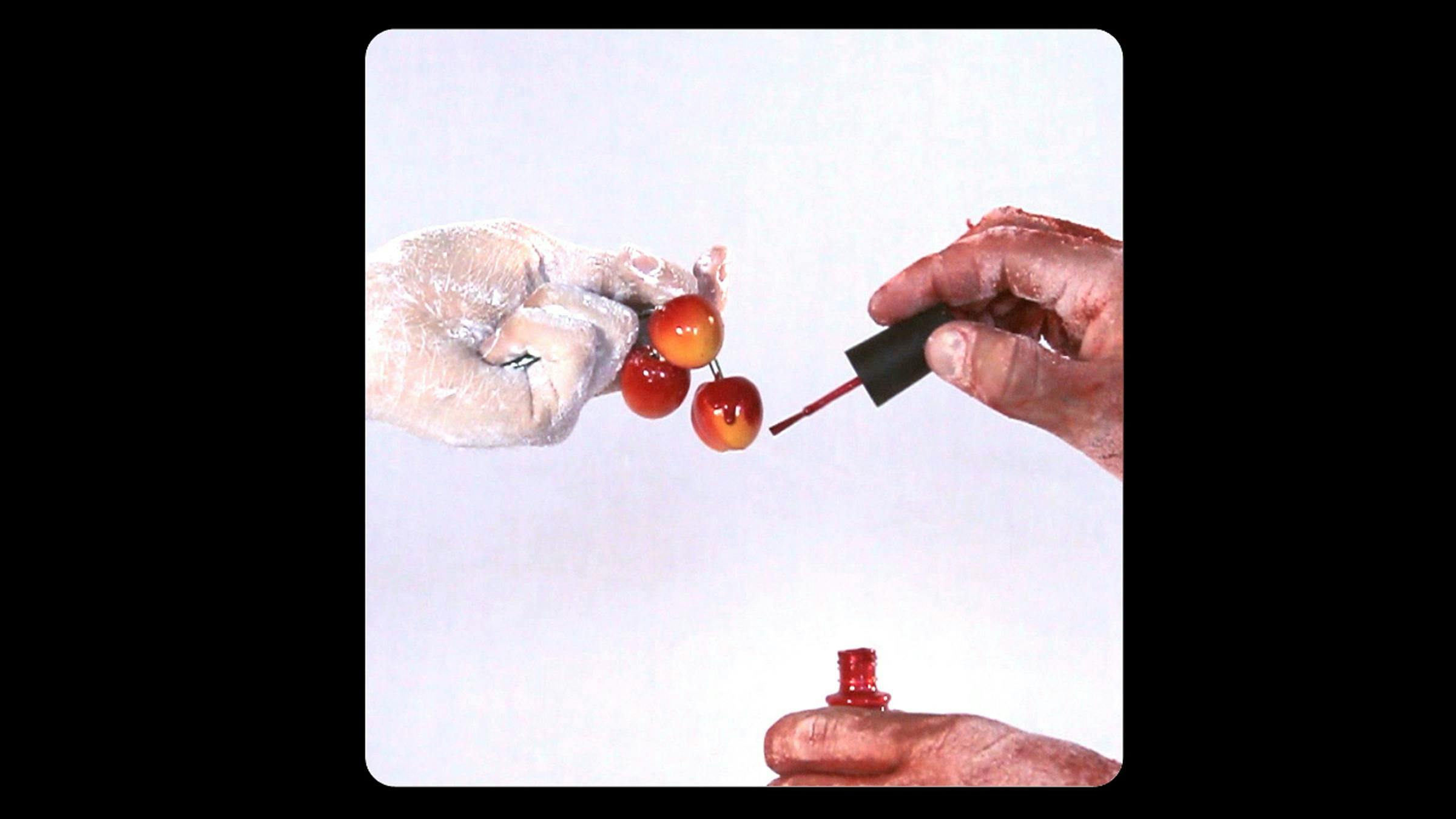 Hand covered in white powder holds three small balls. Another pair of hands covered in red paints the balls with red nail polish.
