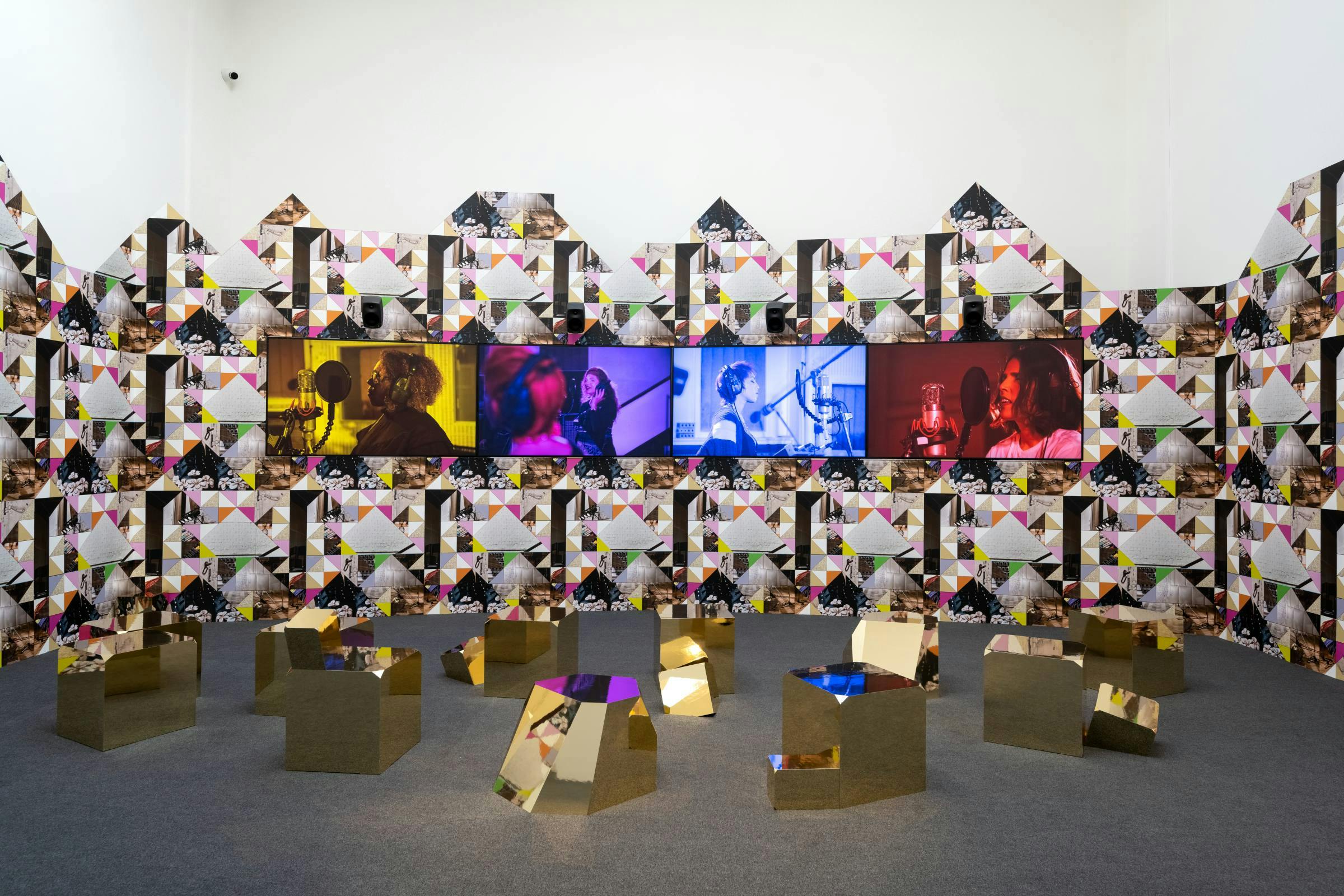 an installation of video screens and gold sculptures in a gallery space. The white wall that is visible is pasted with angular patterned wall paper.