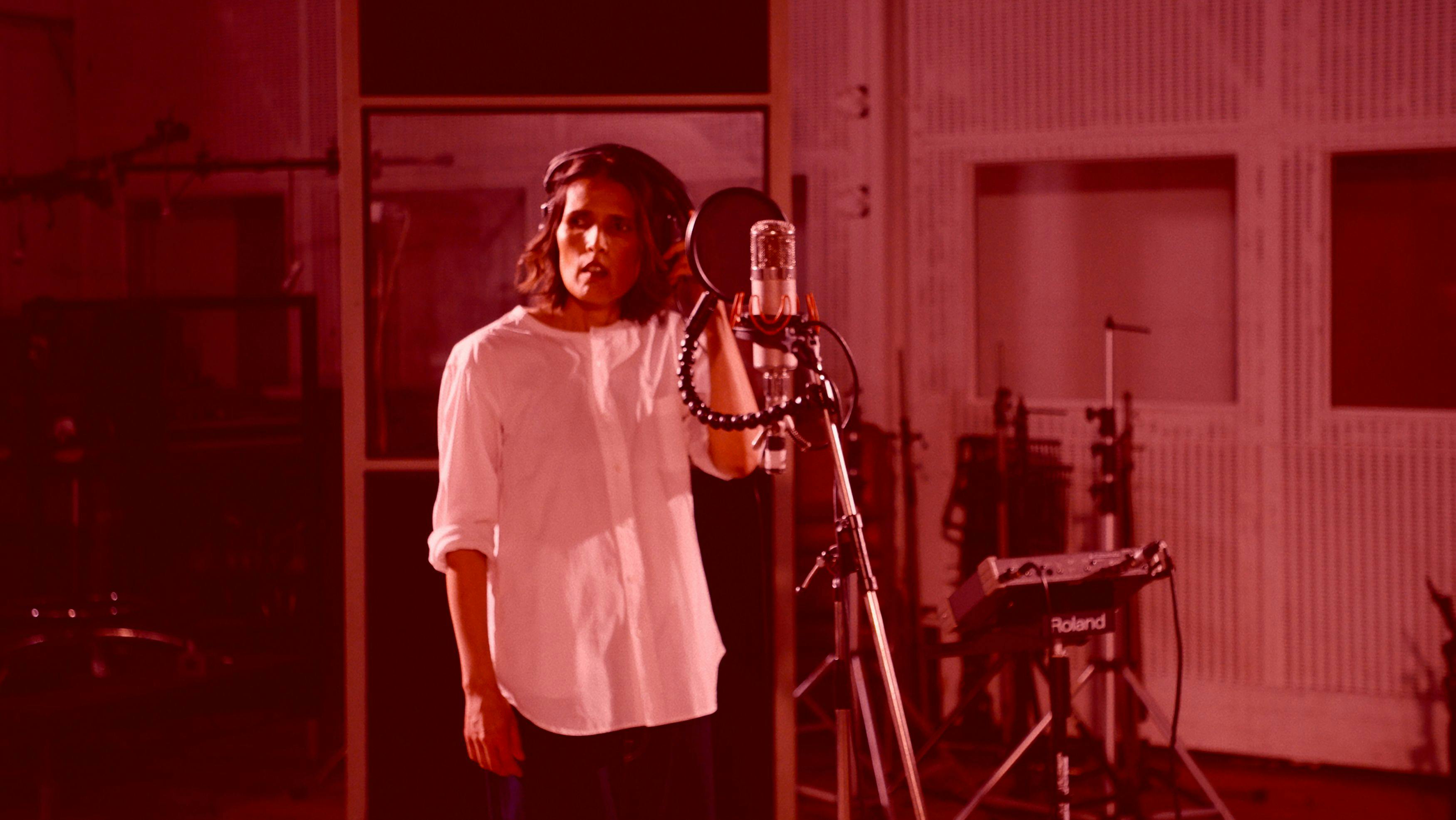 A woman wearing a white shirt is singing into a microphone in a recording studio, the image has a dark pink hue
