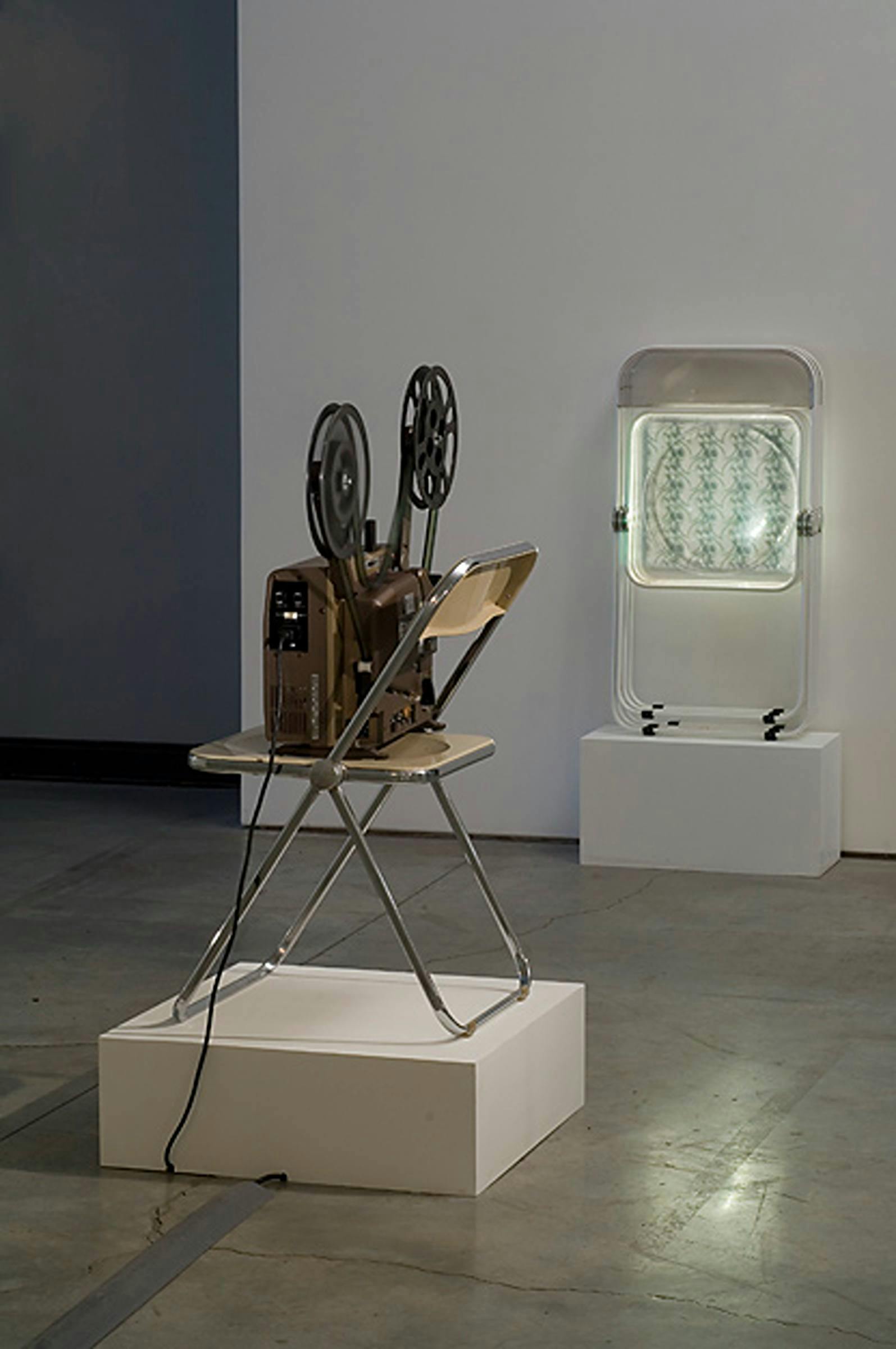Image of a video camera with the film wound at the top, sat on top of a chair on a low-lying plinth. There is an object lent against the other wall also on a low-lying plinth.