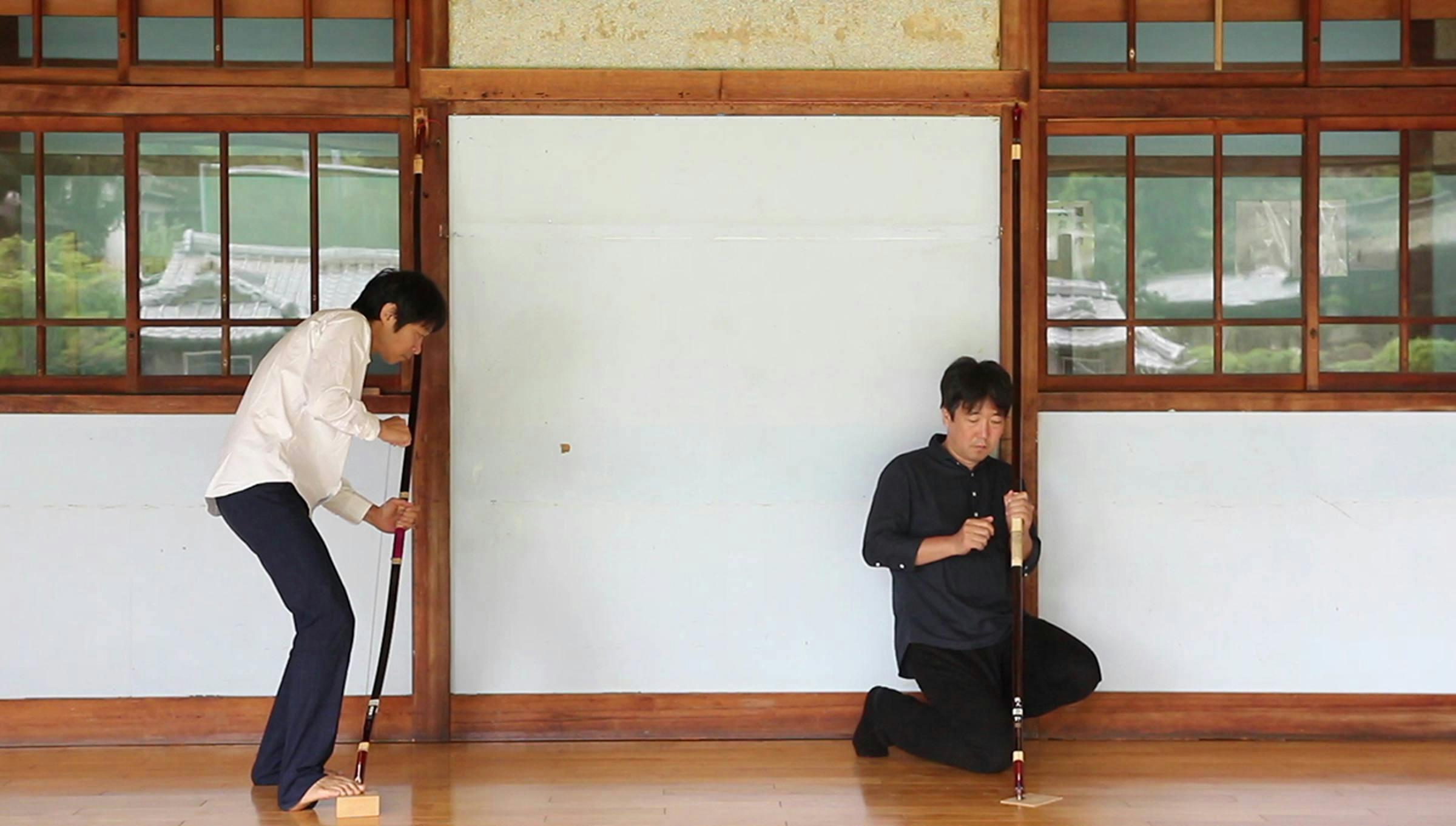 Two people are in a space with a wooden floor. Each person holds an archery bow upright with one end on the floor and they appear to play the bow like an instrument.