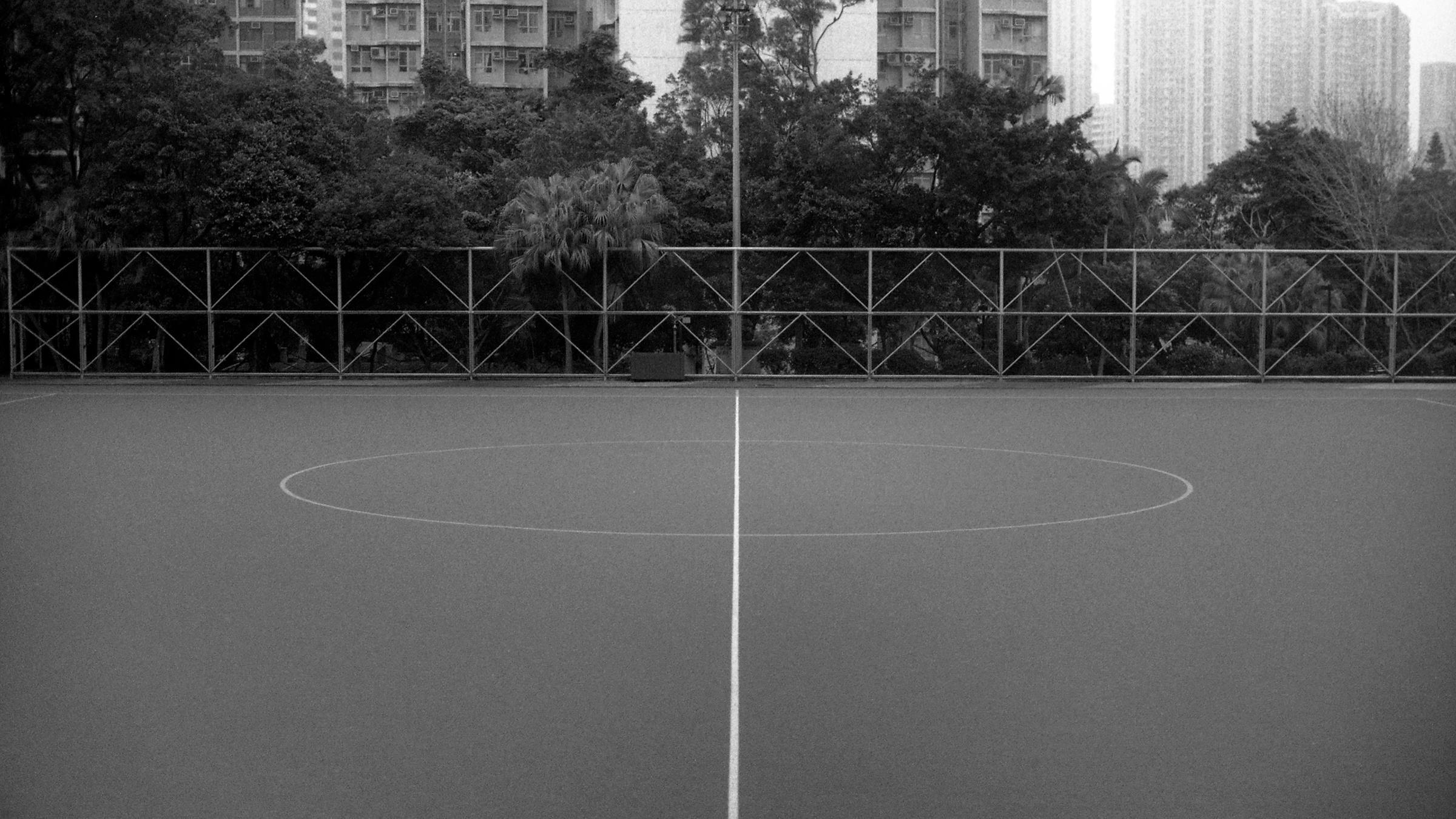Black and white image of a sports court with white lines on the floor. In the background, there are trees and some apartment buildings.