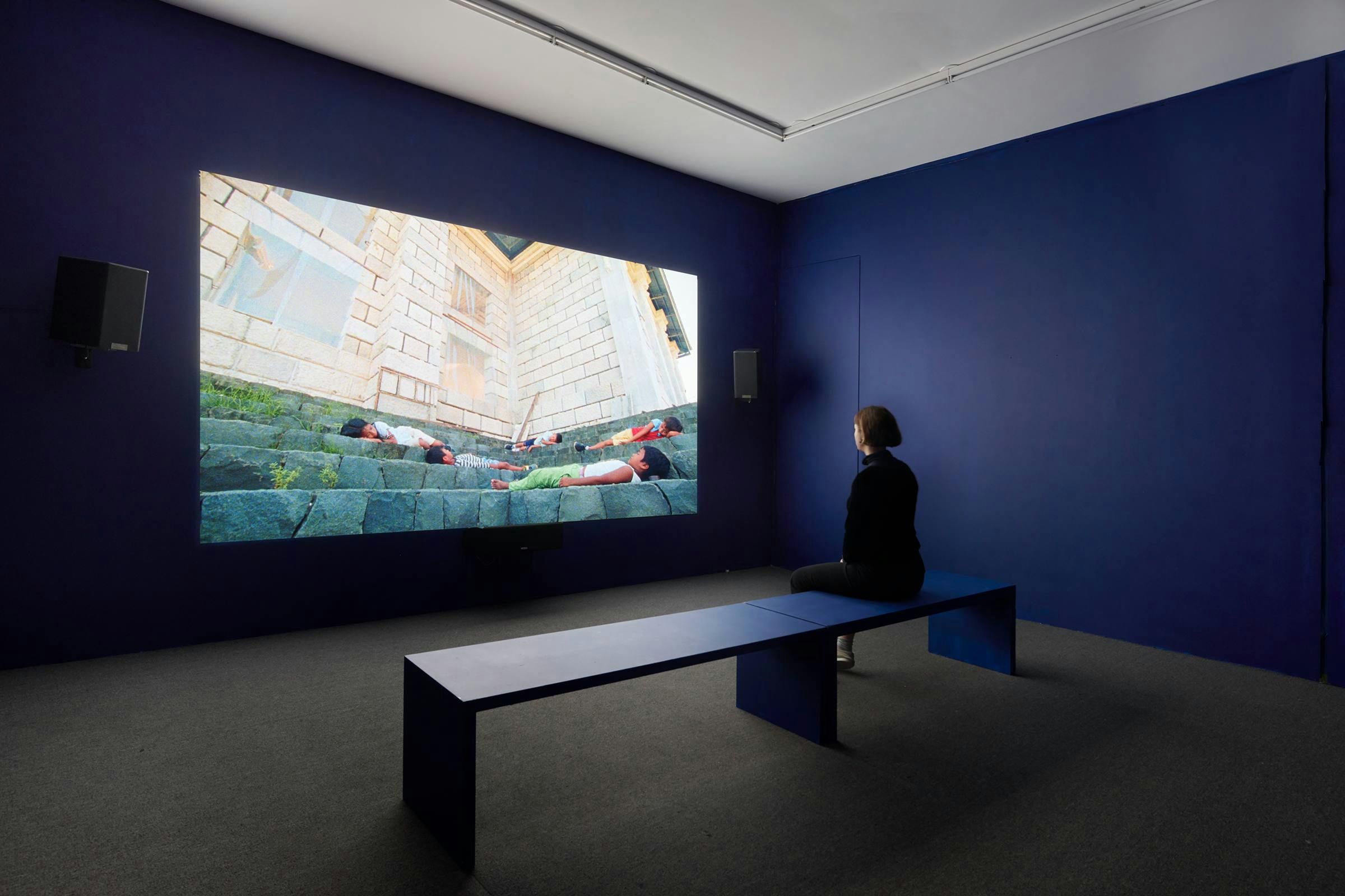 An art work projected in an gallery space. The walls are blue and there is a blue bench in front of the work which a person is sitting on.