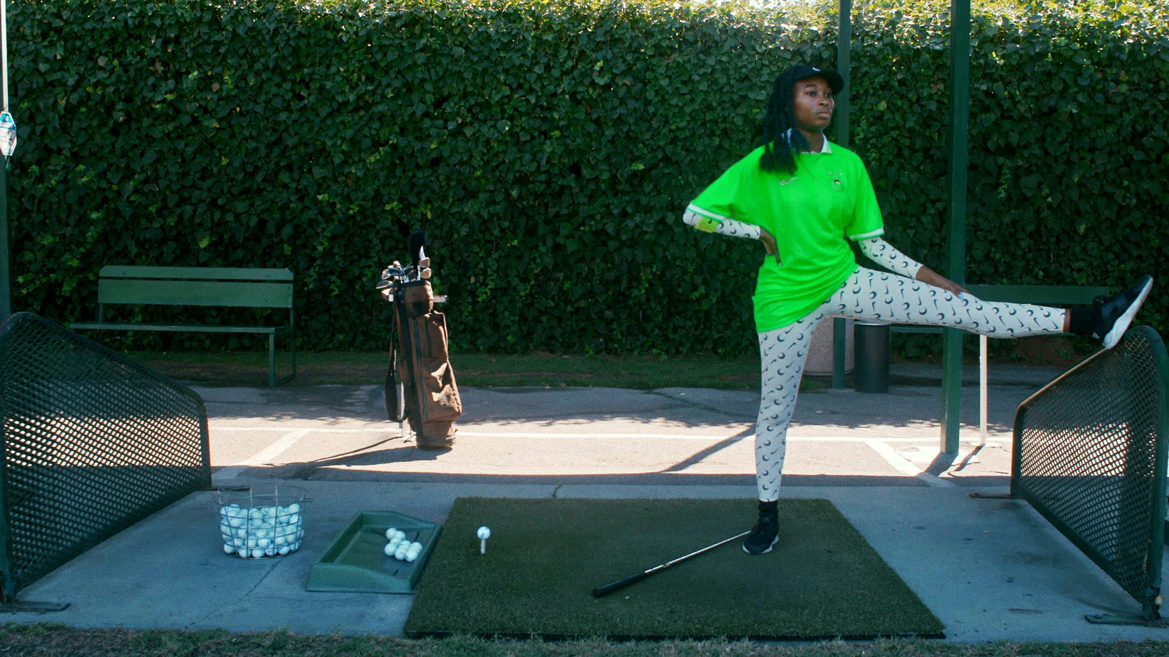 The artist is standing with a stretched leg in a golf practicing course. She is wearing a green jumper and patterned leggings. The sun is shining.