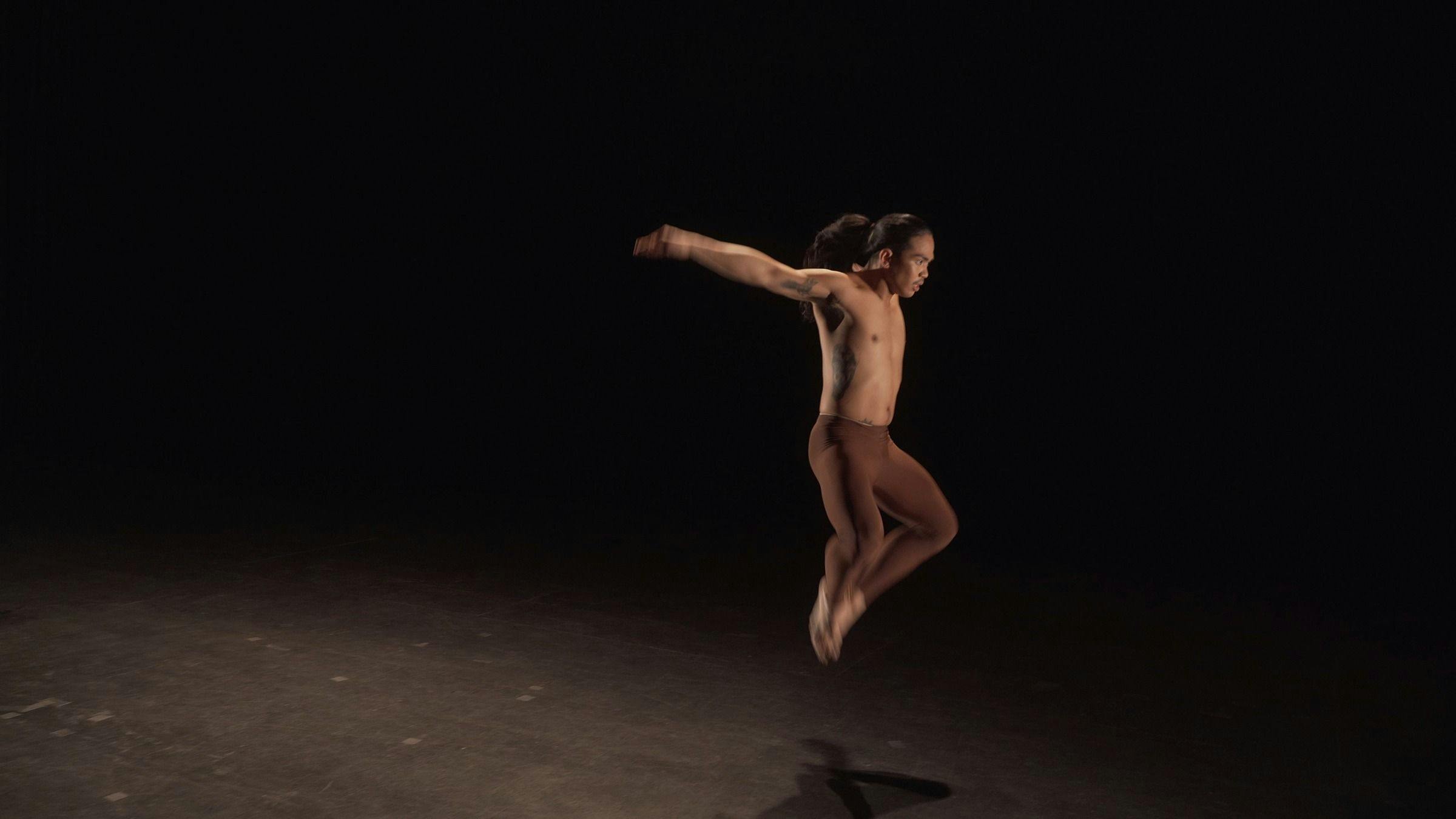 a dancer who is wearing brown leggings leaps in the air in a darkened space