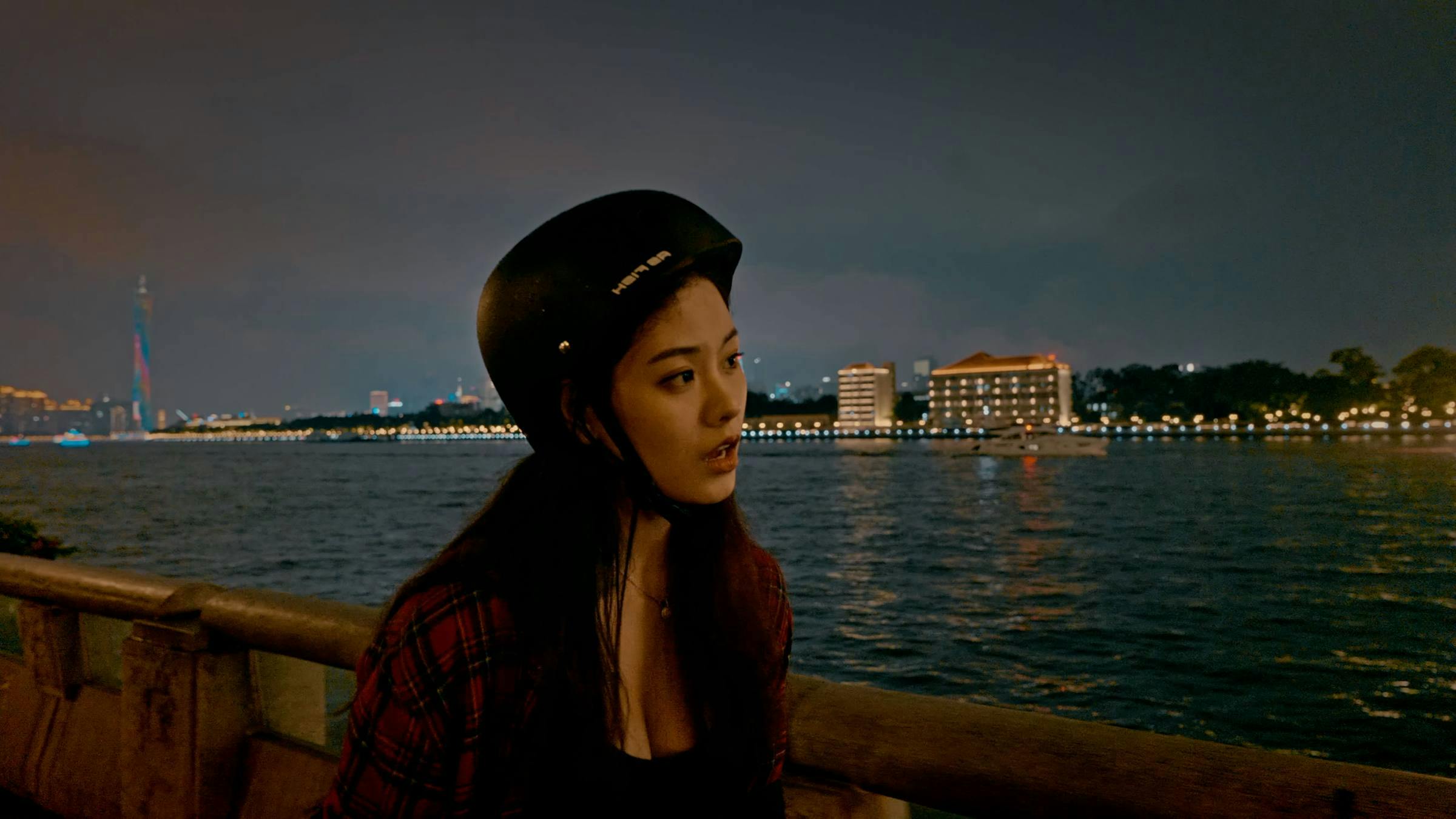 Image of a person wearing a black scooter helmet, stood in front of a fence with water behind them. The image is at night and there are lots of lights from nearby buildings reflecting on the water.