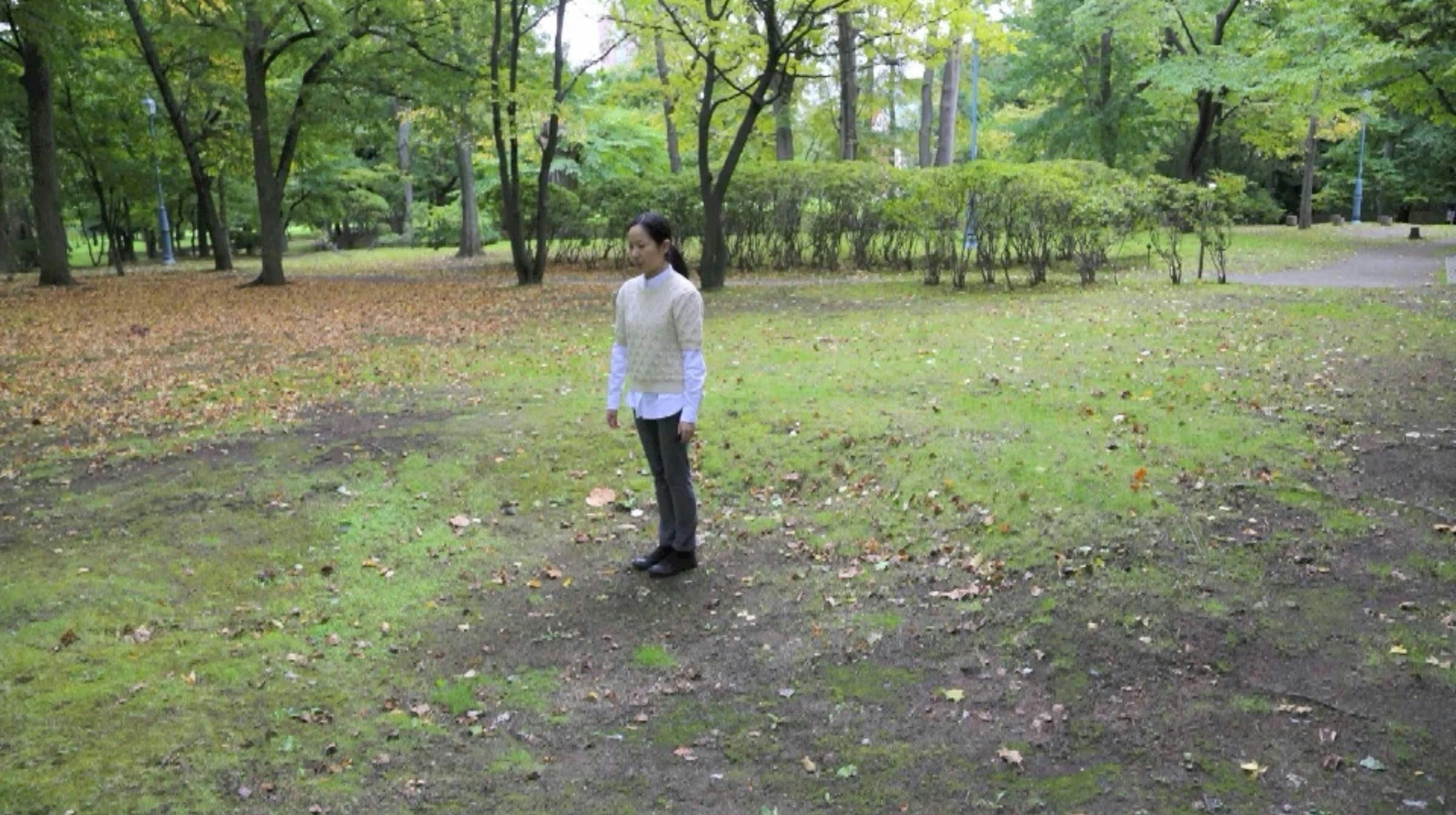 A person stands with their arms down by their sides in a grassy area with trees around them. They are wearing a white shirt with a cream t-shirt on top.