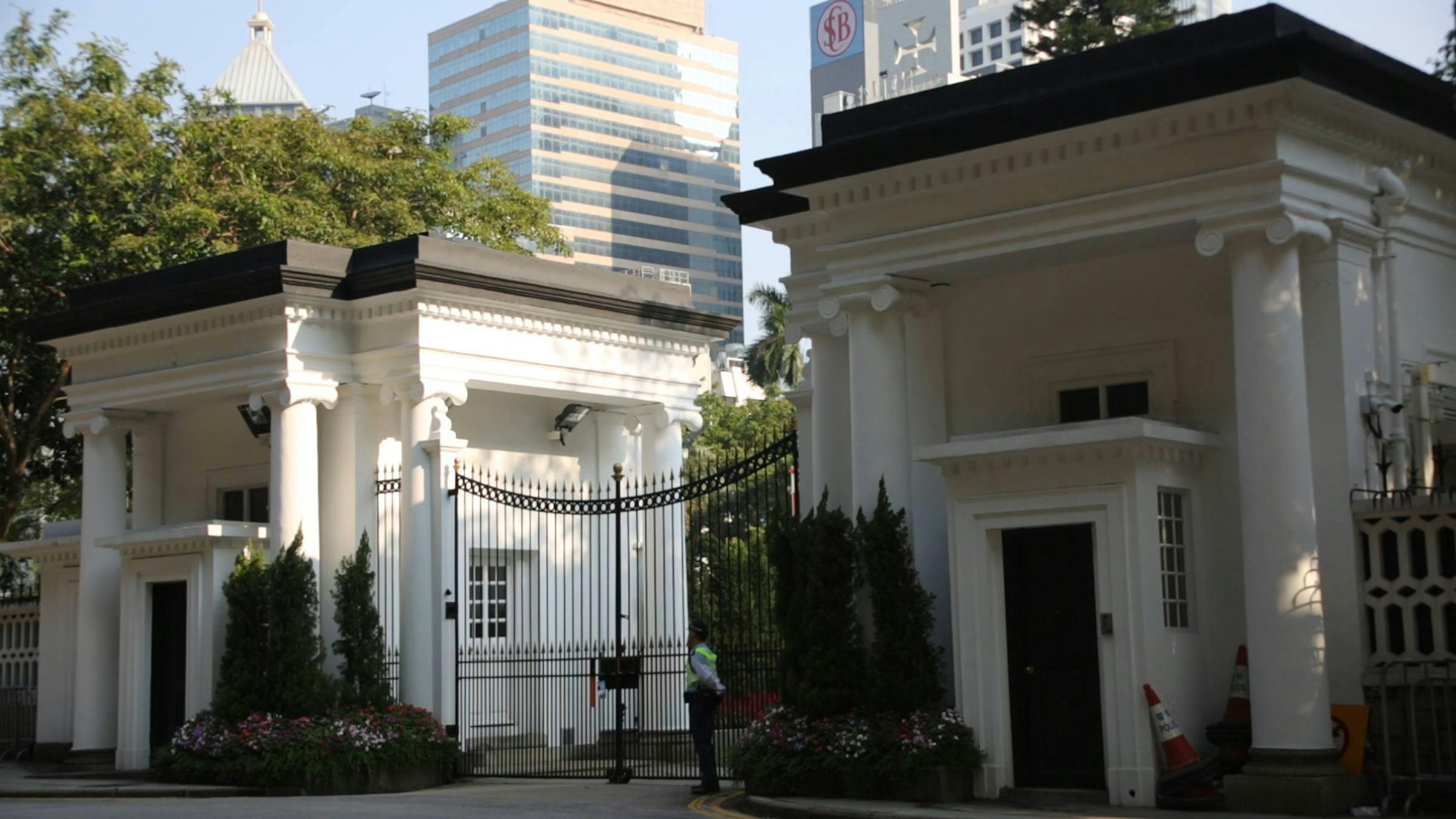 image of two official looking white buildings with columns on every side, with a tall gate in between them. There is a security guard standing next to the gate.