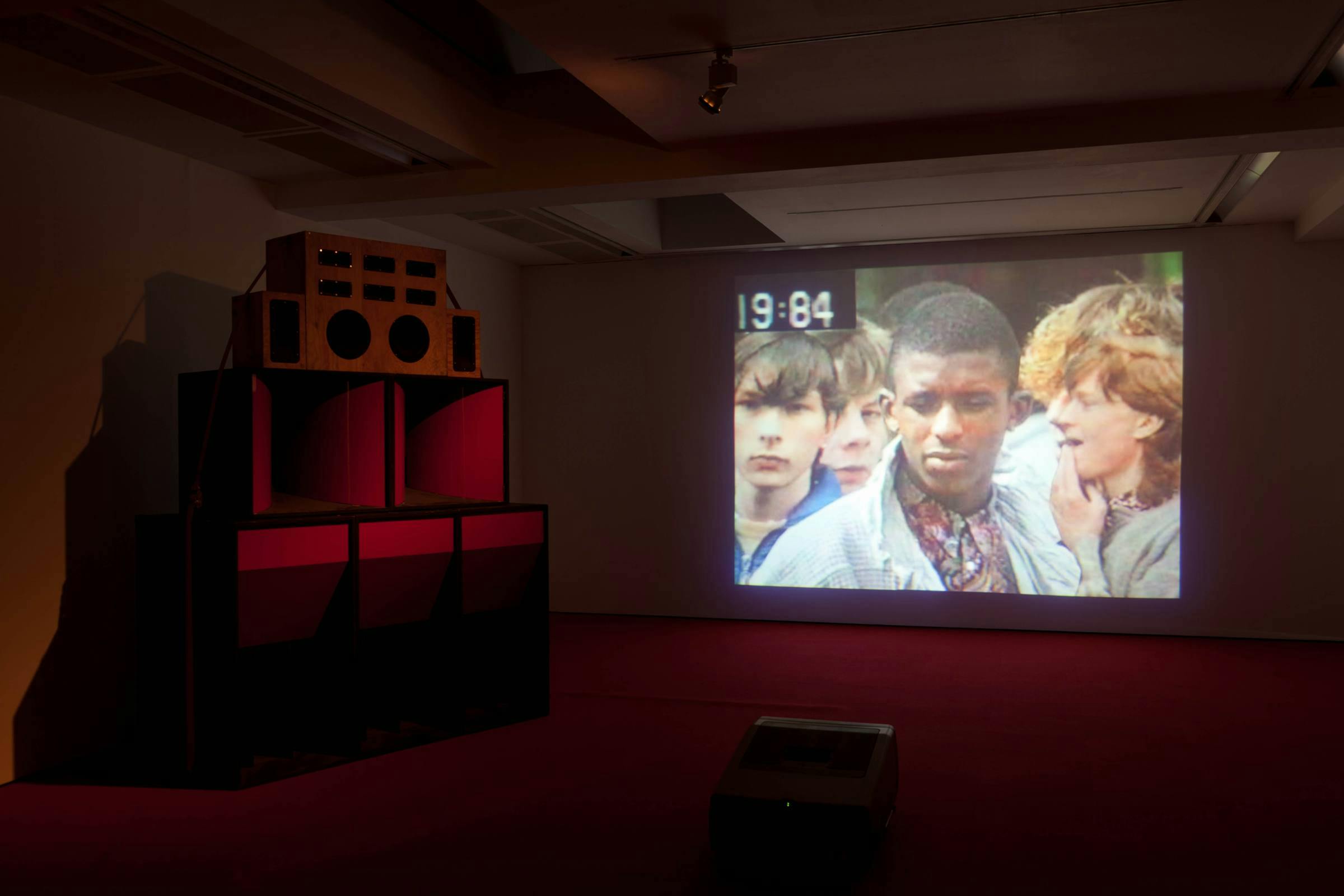 Image of a film playing in a darkened gallery space, the film projected onto one wall with a large speaker-looking structure next to it