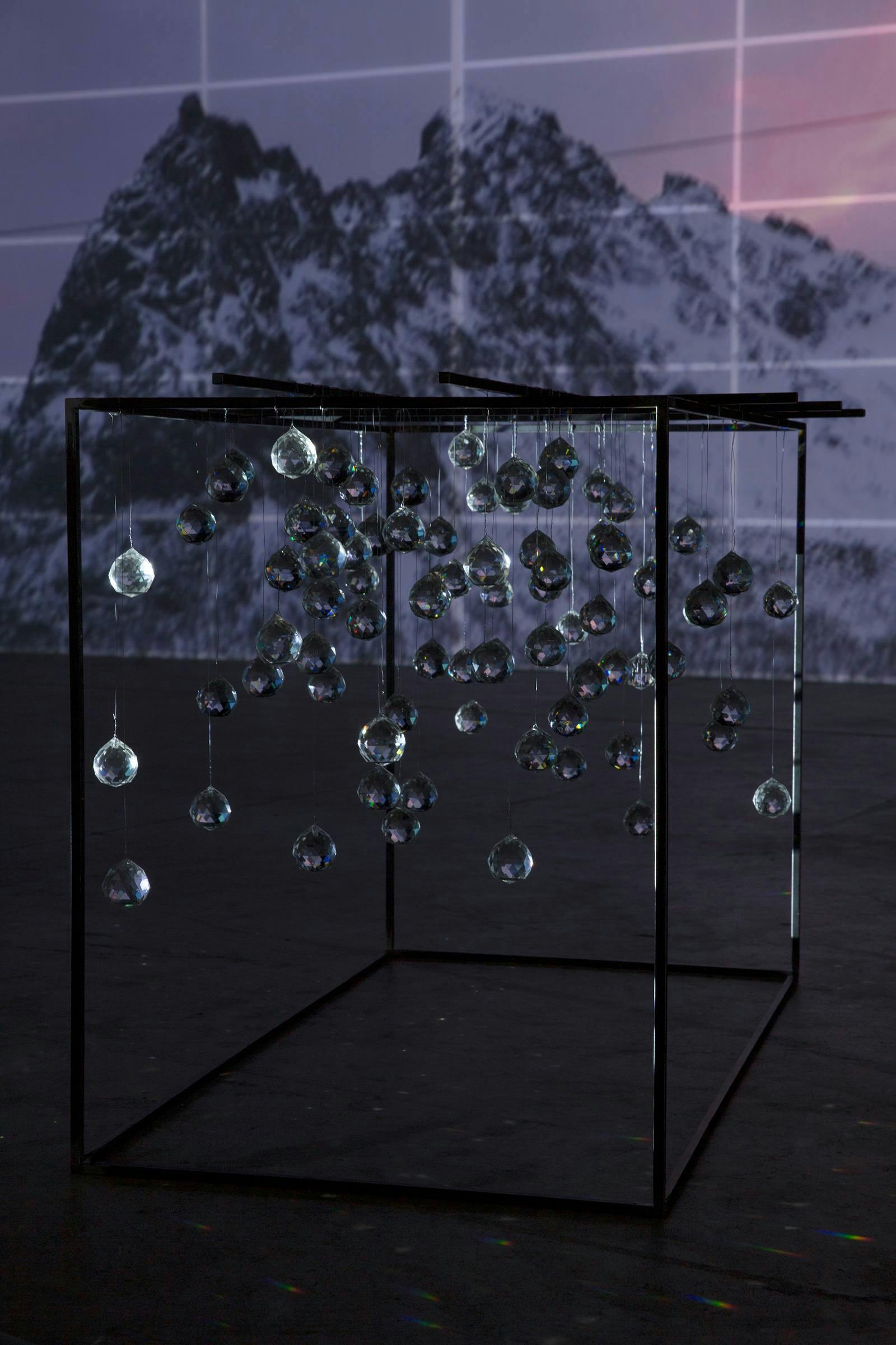 an installation view of Joan Jonas, Ice Drawing Sculpture. There is a projection of mountains in the background and a metal open cube structure with glass spheres hanging from it in the foreground