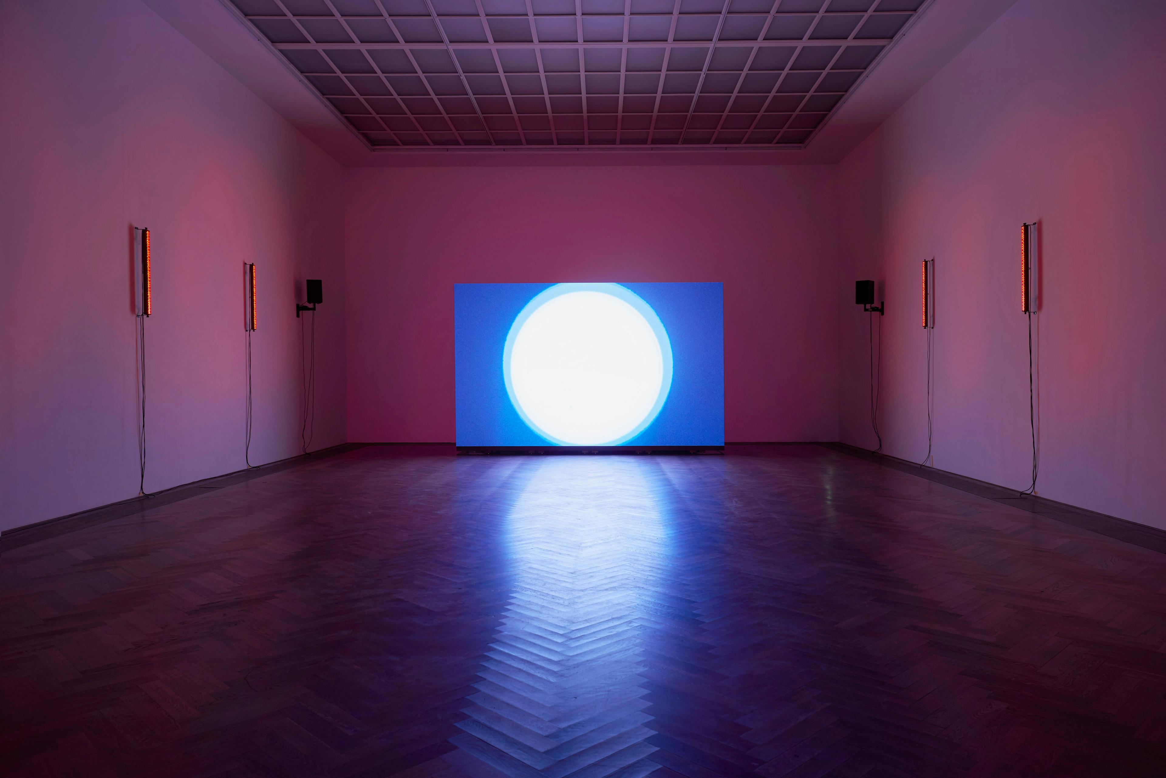 A gallery space with pink glowing light. On either side, red neon lights are fixed to the walls. In the centre, a large canvas shows a white glowing ball with blue around it.