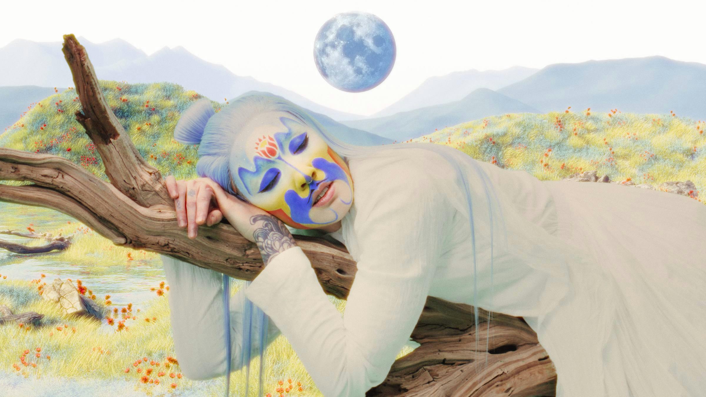 Person wearing white clothing with pale blue long hair and their face painted in blue, orange and yellow, lying on a tree branch