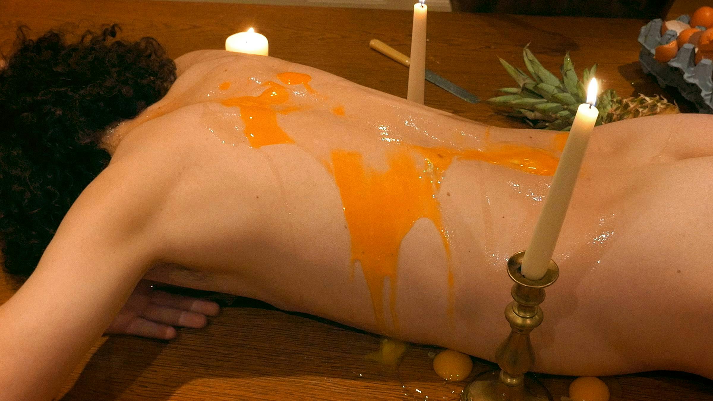 A person lies naked on their front with their head down. Their back is covered in a yellow liquid and raw eggs are by their sides. There are candles around them and a pineapple.