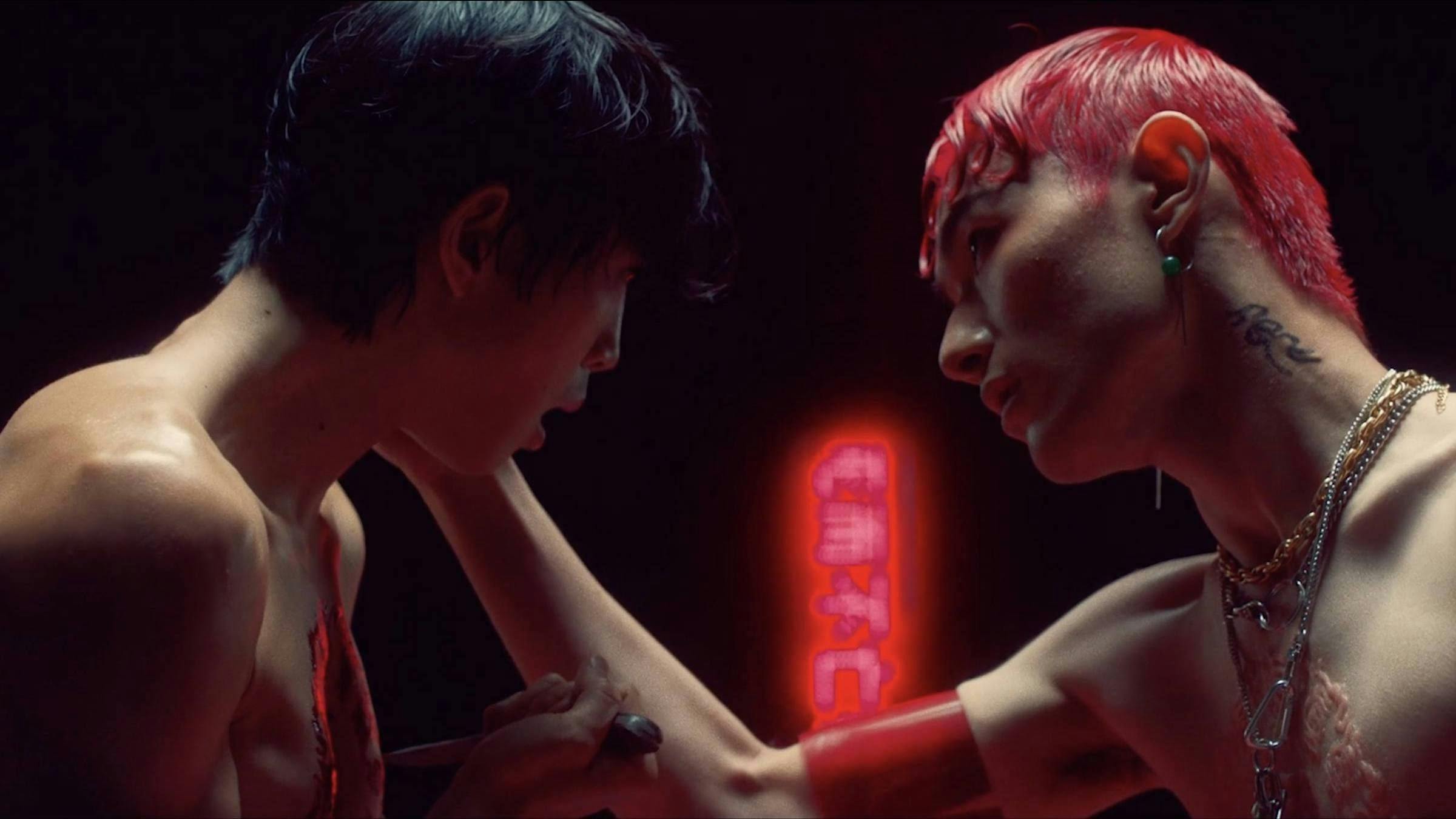 2 shirtless people face each other and seem to be leaning into each other to kiss. One person has red hair the other has black. We can see Chinese language characters in the background emanating red light. The space is dark