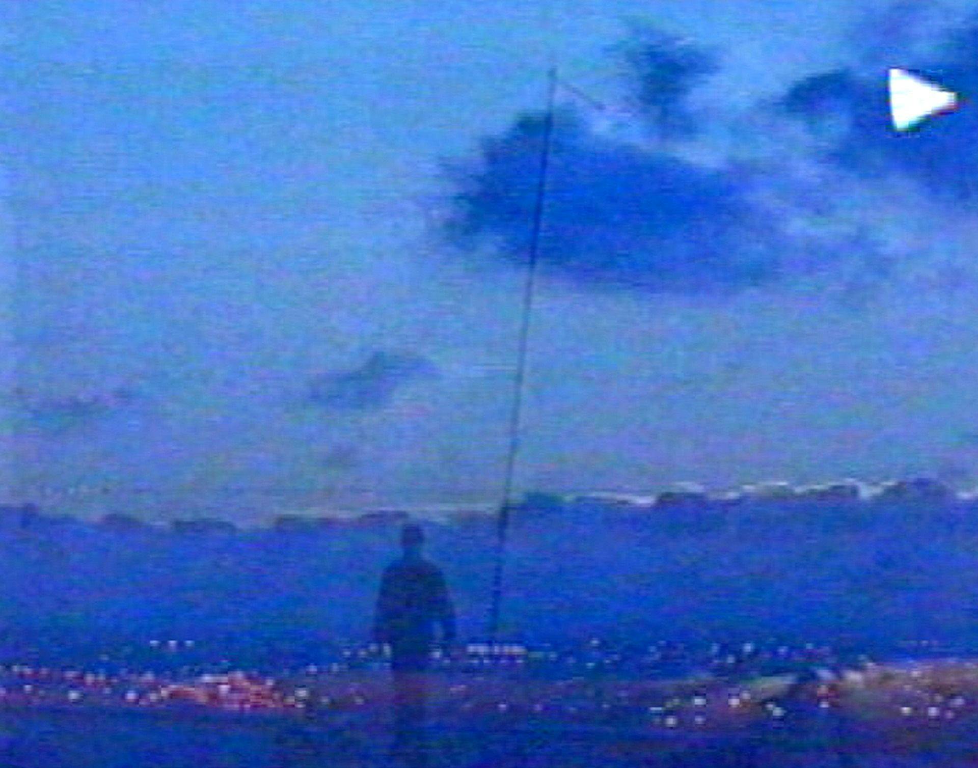 Image of hazy figure in the foreground with city lights on the horizon and a partly cloudy sky above. A play sign appears in the top right corner, as if its an image of a VHS player.