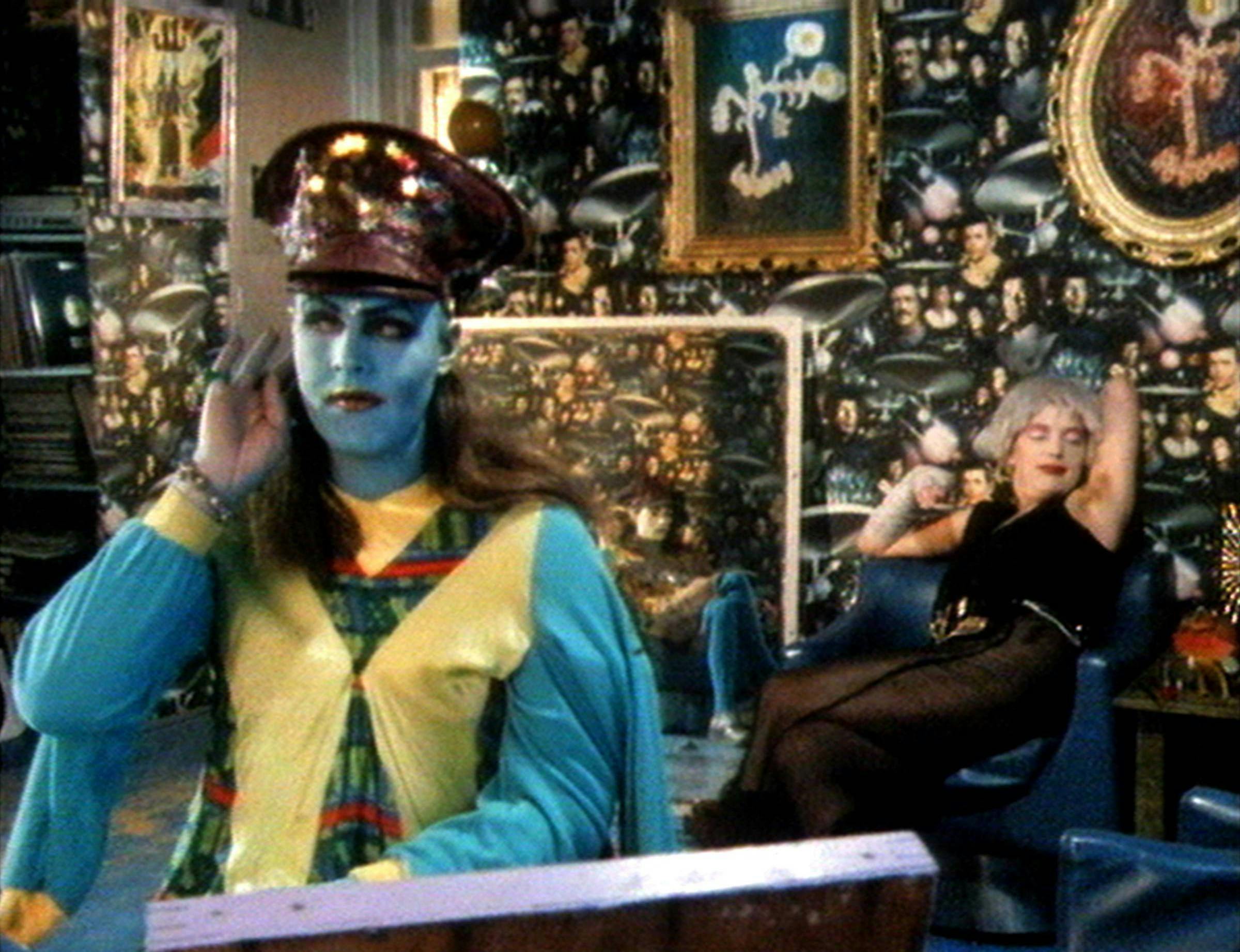Someone sits in the foreground holding their hand up to their ear. Their face is painted blue and they're wearing a large hat. In the background, someone sits reclining backwards. The room is decorated with a floral wallpaper.