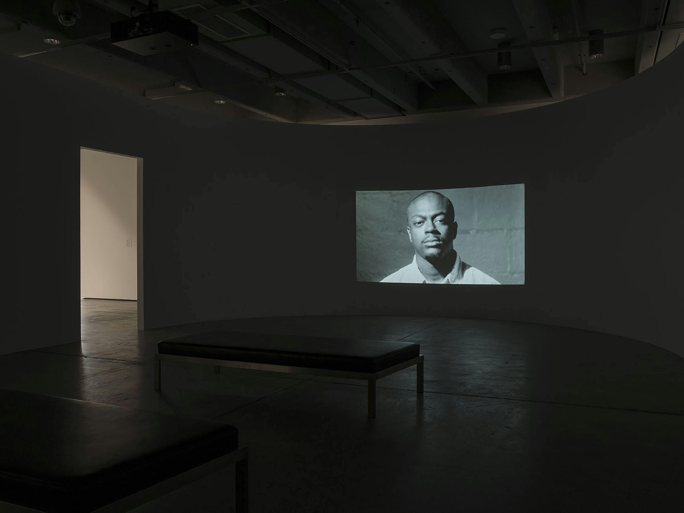 Image of an empty gallery space with large projection on one wall, a bench, and an archway.