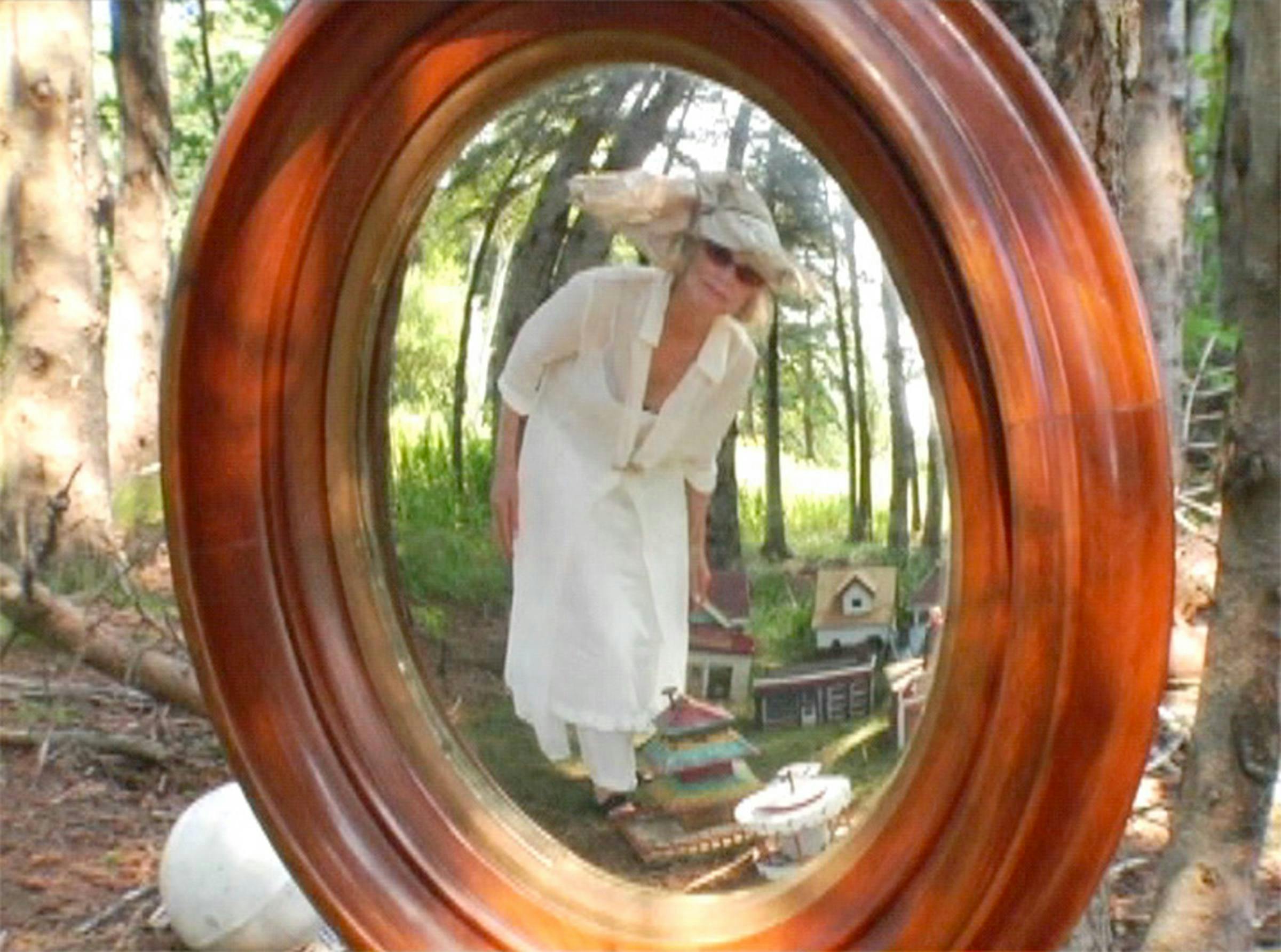 An image of the artist dressed in white and standing in a garden. She is wearing sunglasses. She is peering through a wooden window at us