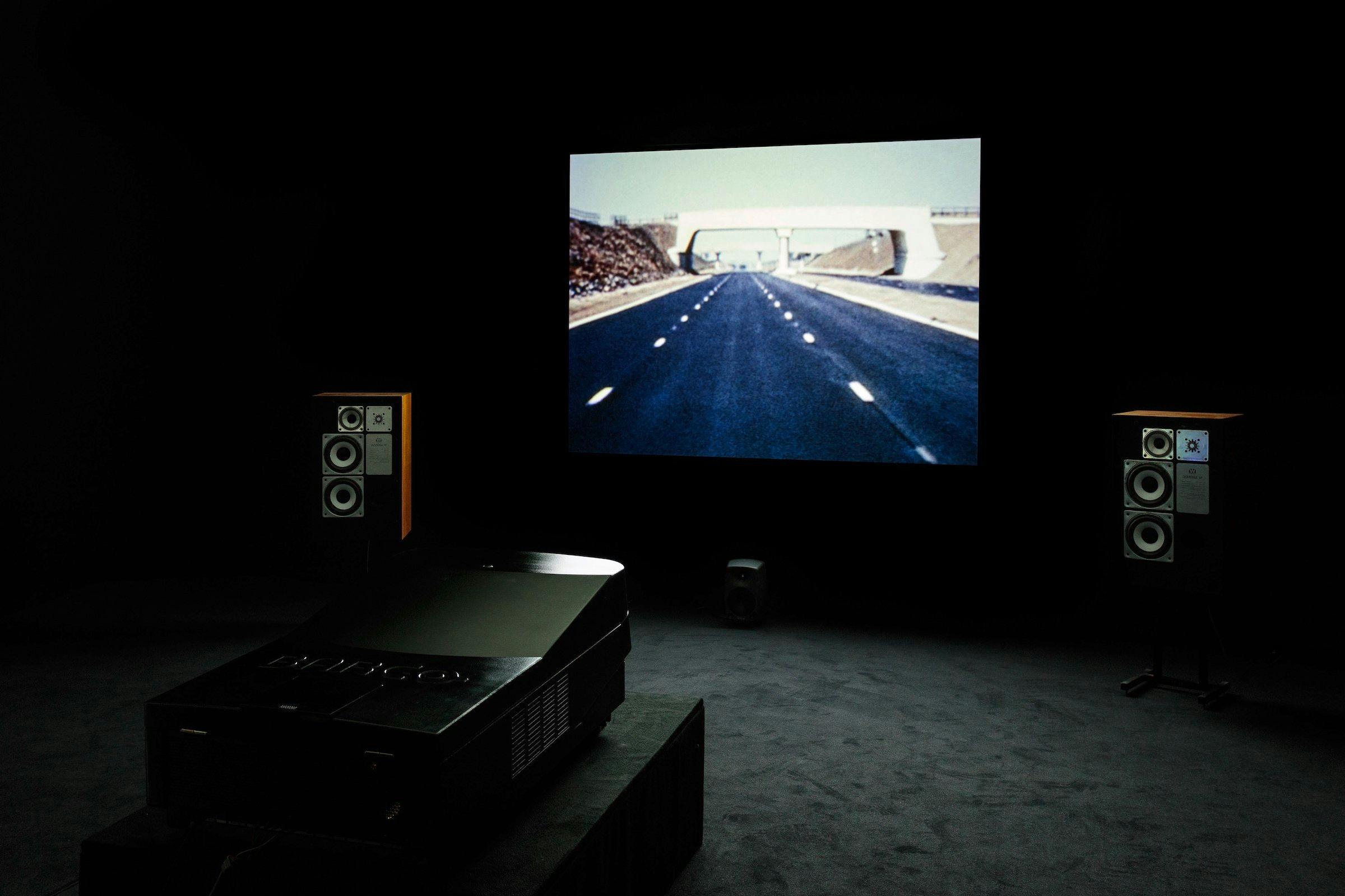 an installation view of Dream English Kid, 1964-1999 AD. The image of the work projected is a POV person driving a car. There are speakers either side of the projection and the projector is visible
