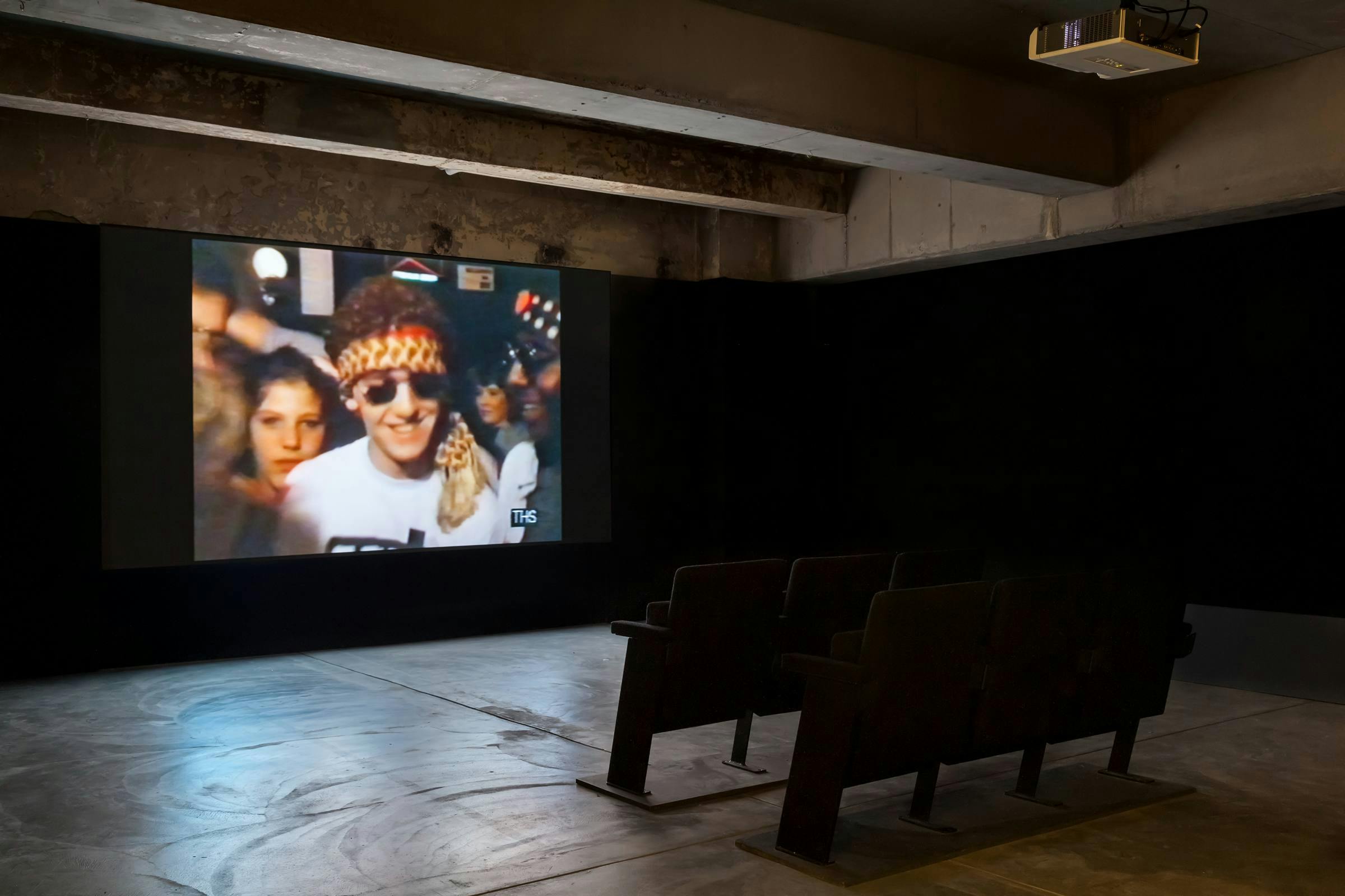 Image of a film playing in a darkened gallery space with two rows of seats and a projector above