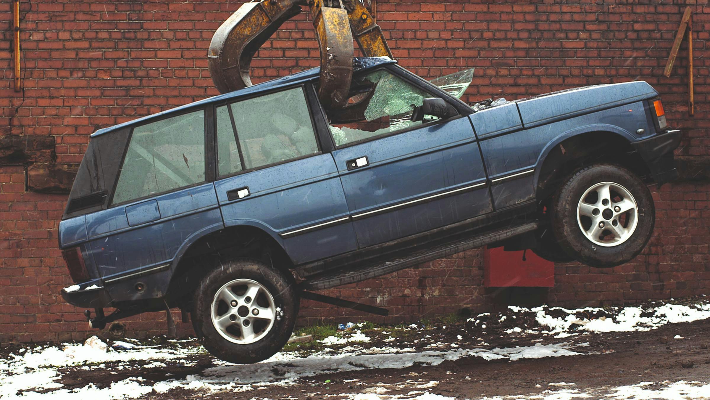 a blue car being lifted by a crane. The forks of the crane have shattered the car's side windows. The ground has snow on it and we can see a red brick wall in the background