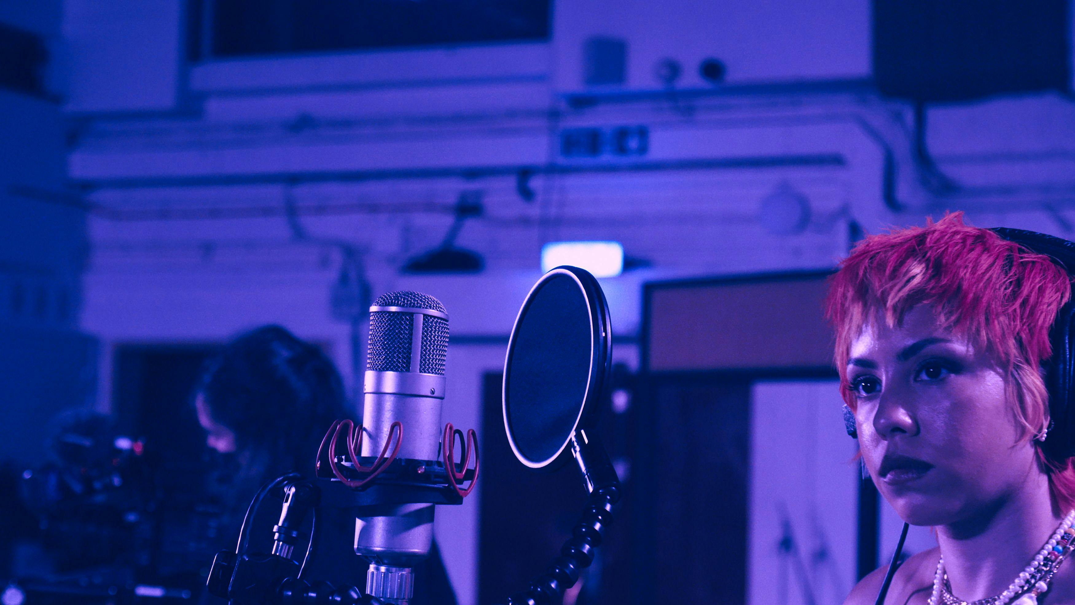 A black woman with red short hair is singing into a microphone in a recording studio, the image has a purple hue