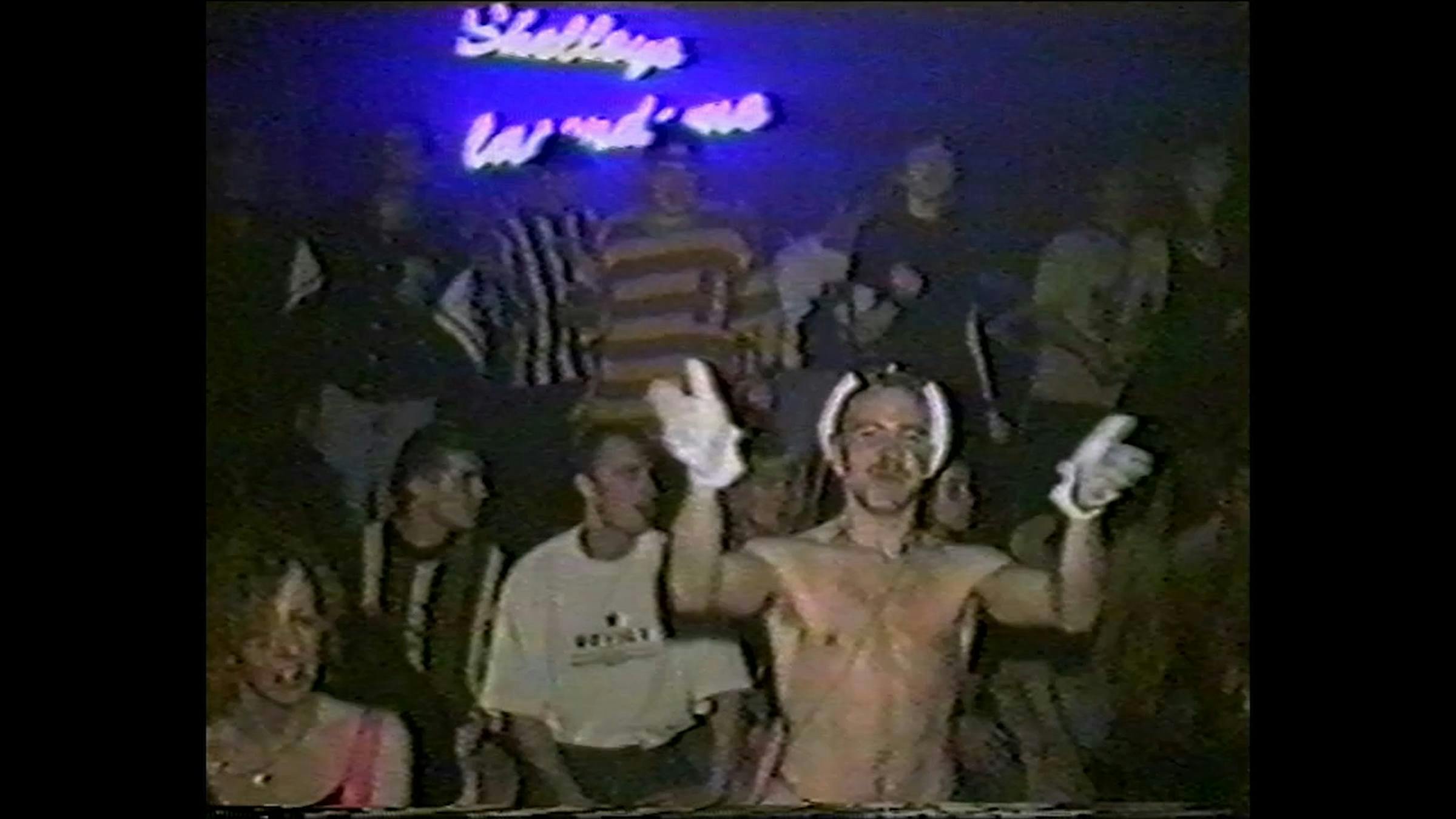 an image of people dancing at a rave in the 1990s. The person in the foreground is shirtless and is wearing white gloves on his hands