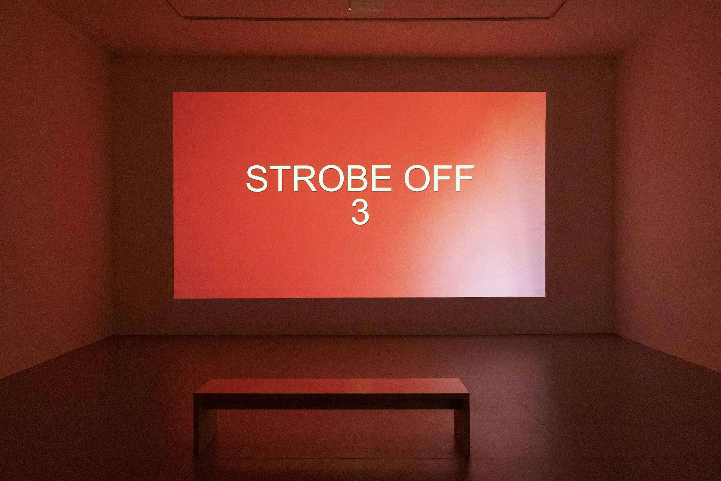 Projection of a red image with words 'STROBE OFF 3' in white projected in a gallery space.