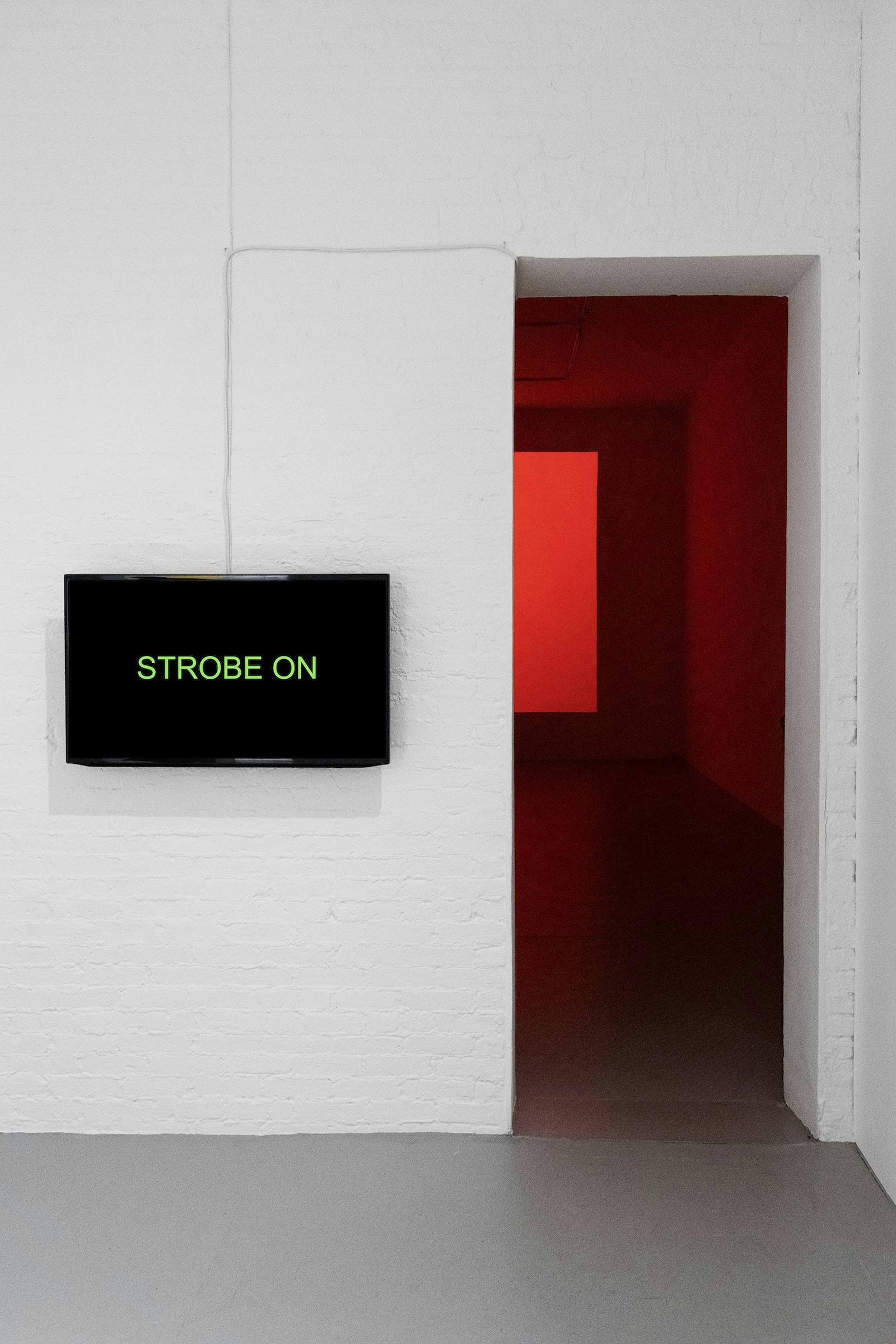 TV screen with black background and 'STROBE ON' in green letters, mounted on a white wall. To the right, there is an archway into a red lit room.