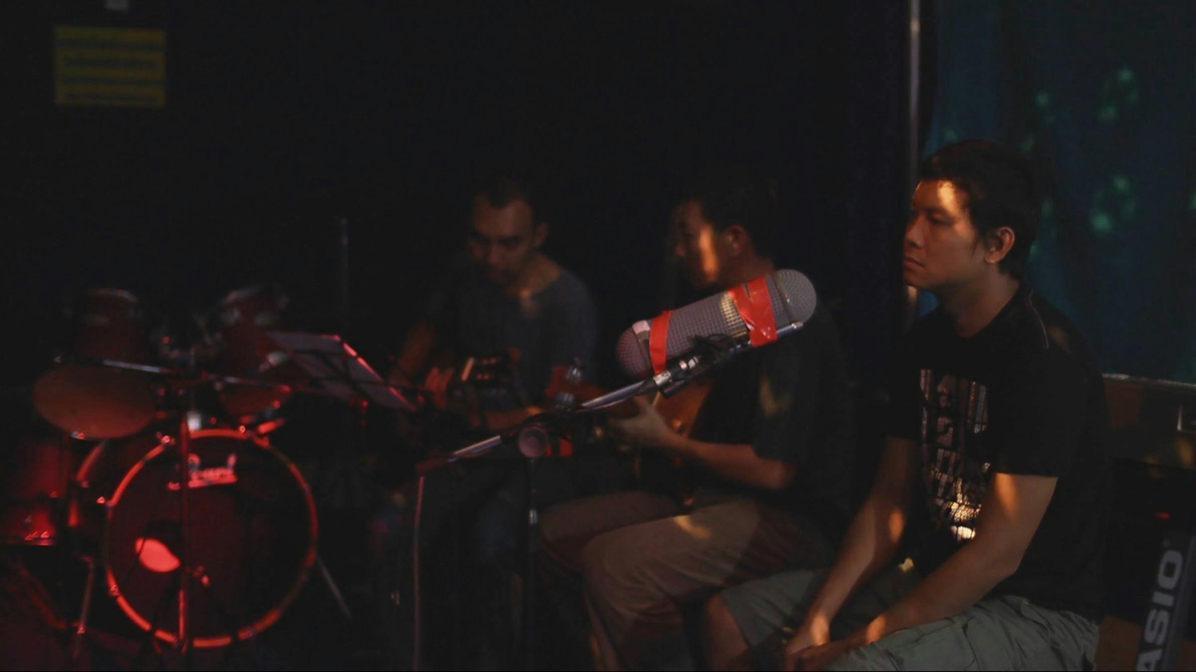the artist is sitting in a room that is green and in front of a very large microphone. There is a person sitting to his right. The room feels as though the people in the image might be playing to a live audience