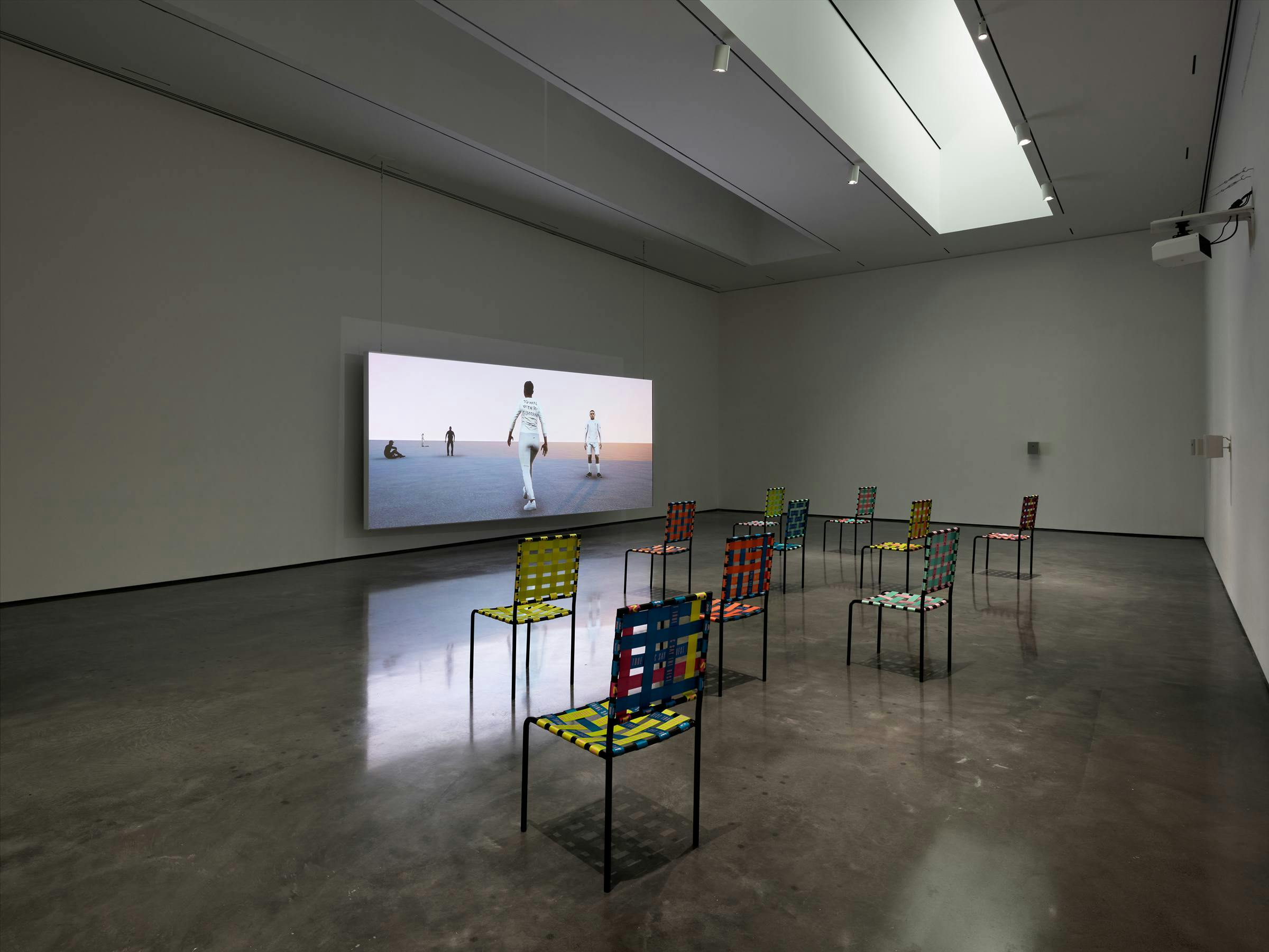 Image of a gallery space with a large projection on one wall with colorful chairs facing it.