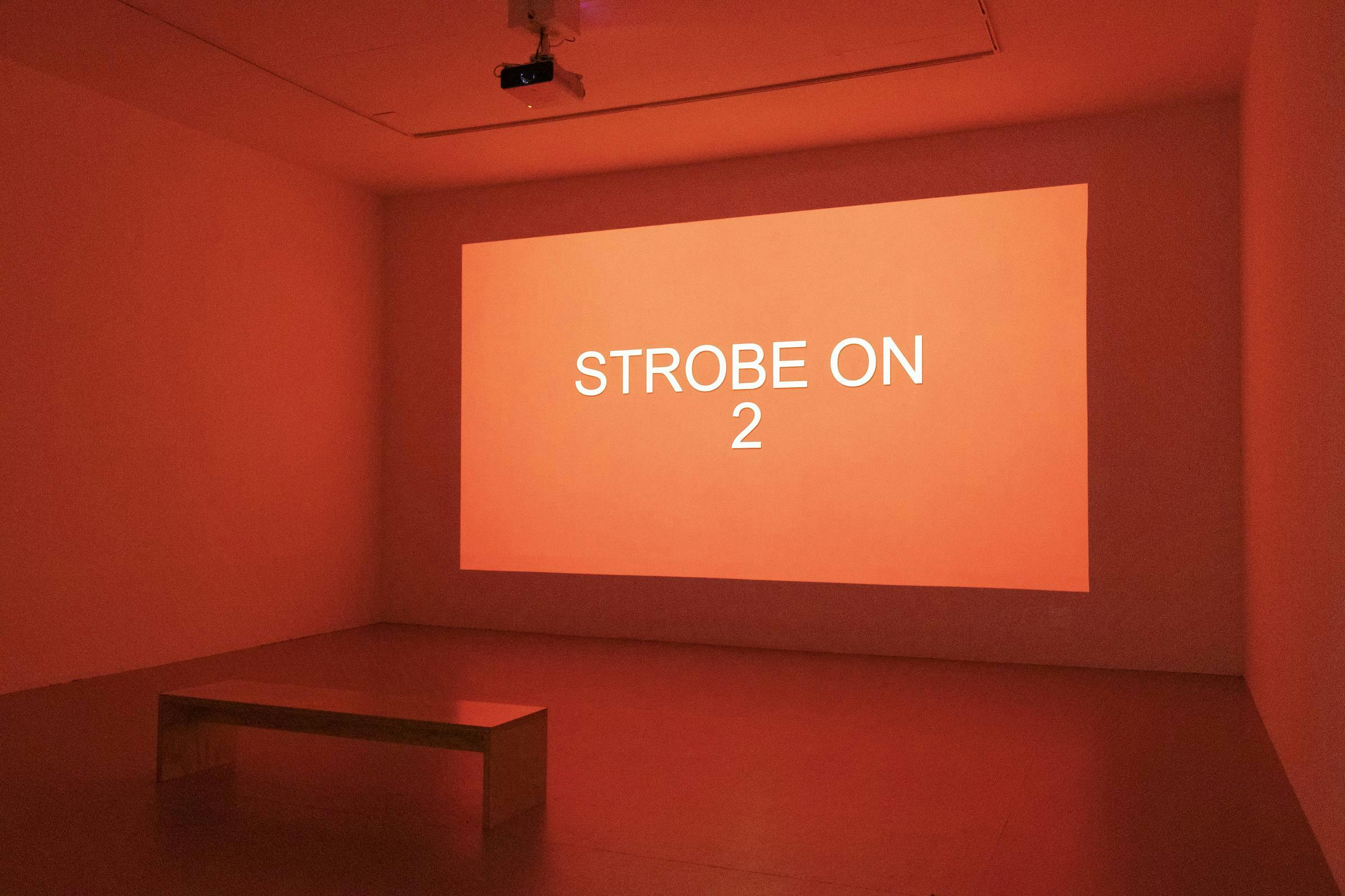 Projection of red background with white writing which reads 'STROBE ON 2' in a gallery space.