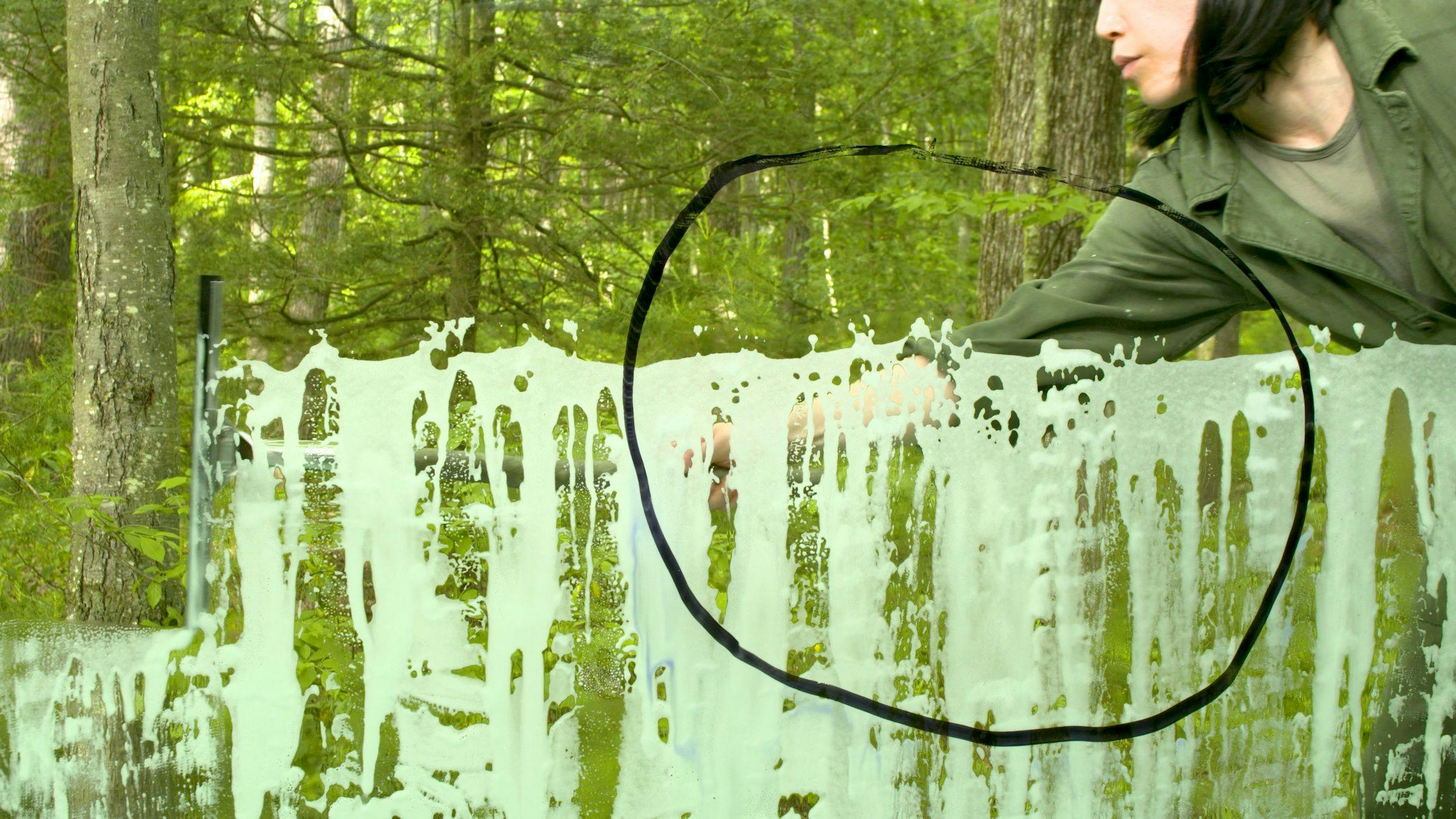the artist has drawn a black circle on the camera lens which is filming a scene in a forest. The artist is wiping soapy suds from the camera lens