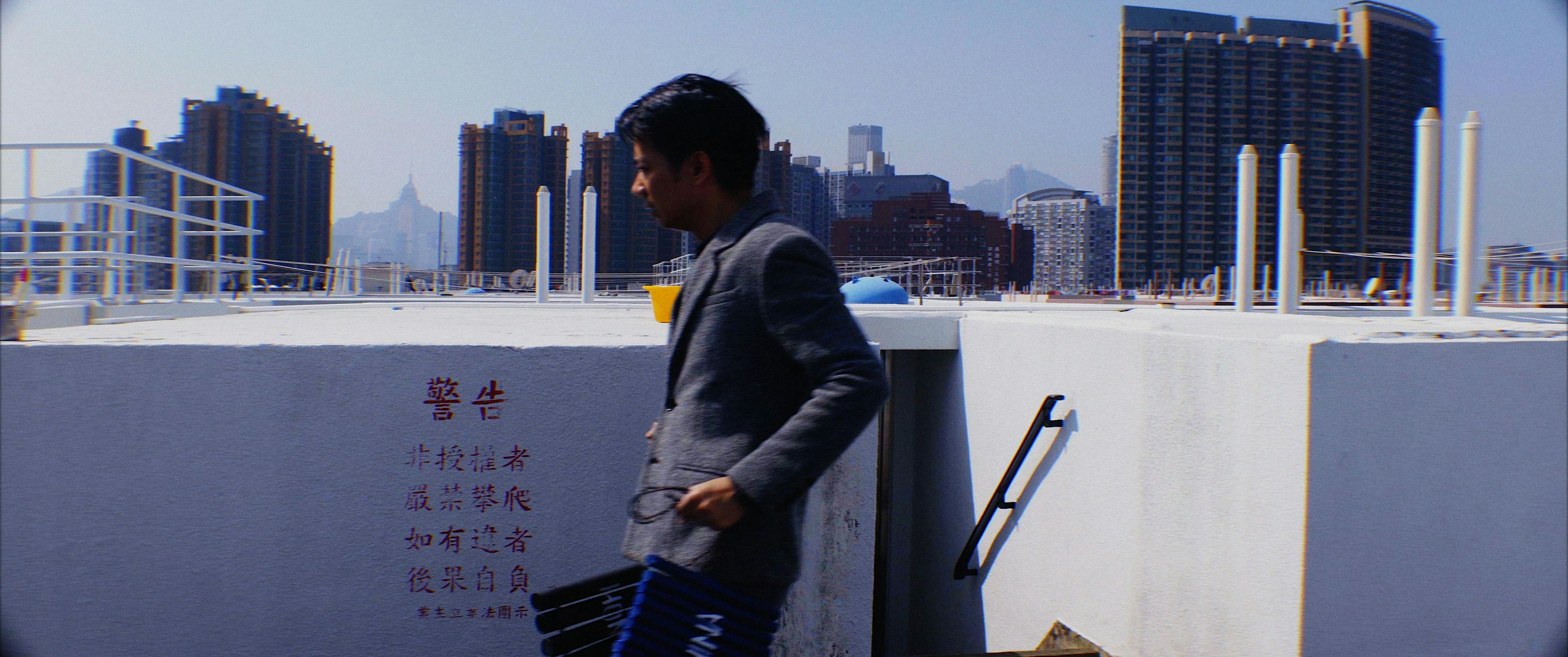Image of a the side profile of a person wearing a grey blazer. They are on the roof of a building in a city and there are skyscrapers in the background.