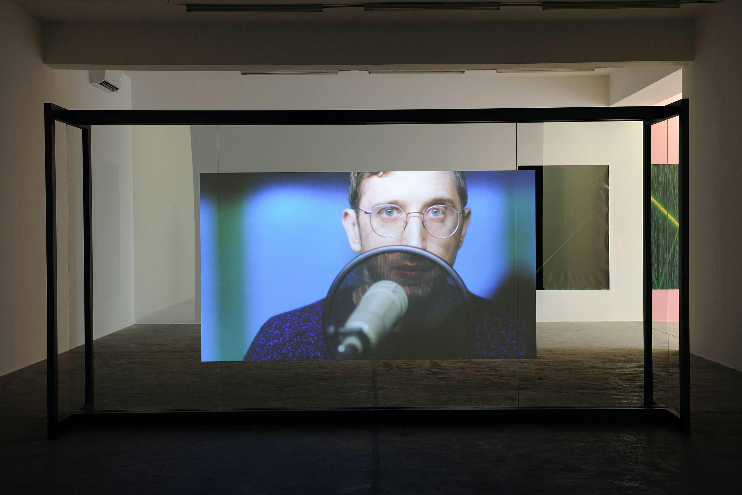 a projection of a video work in a gallery space which shows the artist who is wearing glasses standing in front of a microphone. 