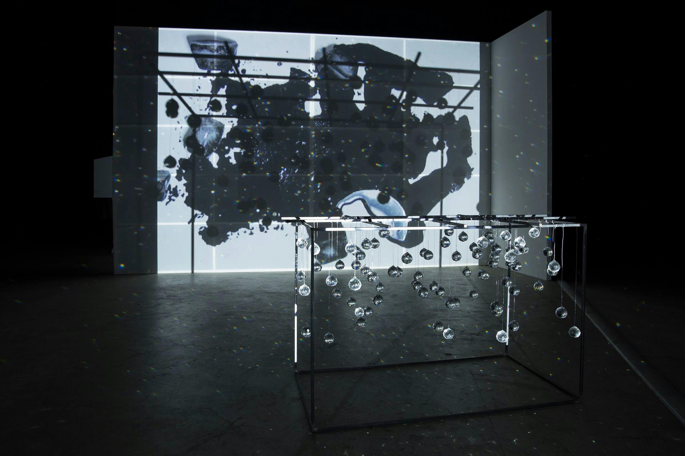 an installation view of Joan Jonas, Ice Drawing Sculpture. There is a projection of melted ice cubes and black ink in the background and a metal open cube structure with glass spheres hanging from it in the foreground