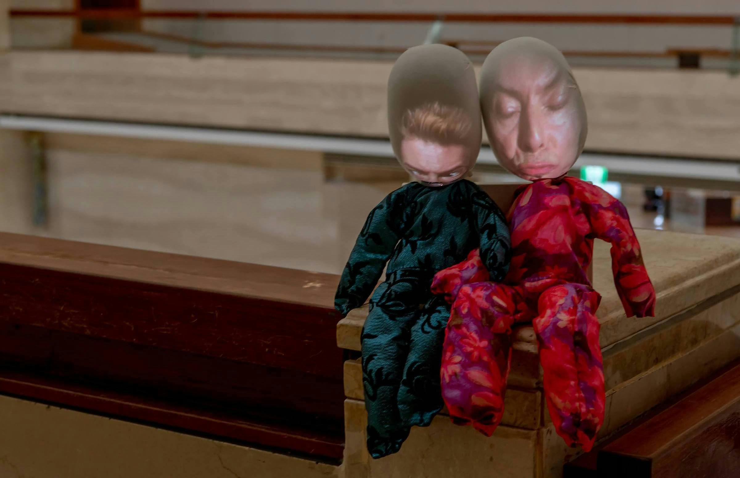 an image of db an art work that comprises 2 dolls. The dolls have the faces of David Bowie projected on to them and the projector is visible. The dolls are installed on the top of a stairwell of a marble and grand building interior