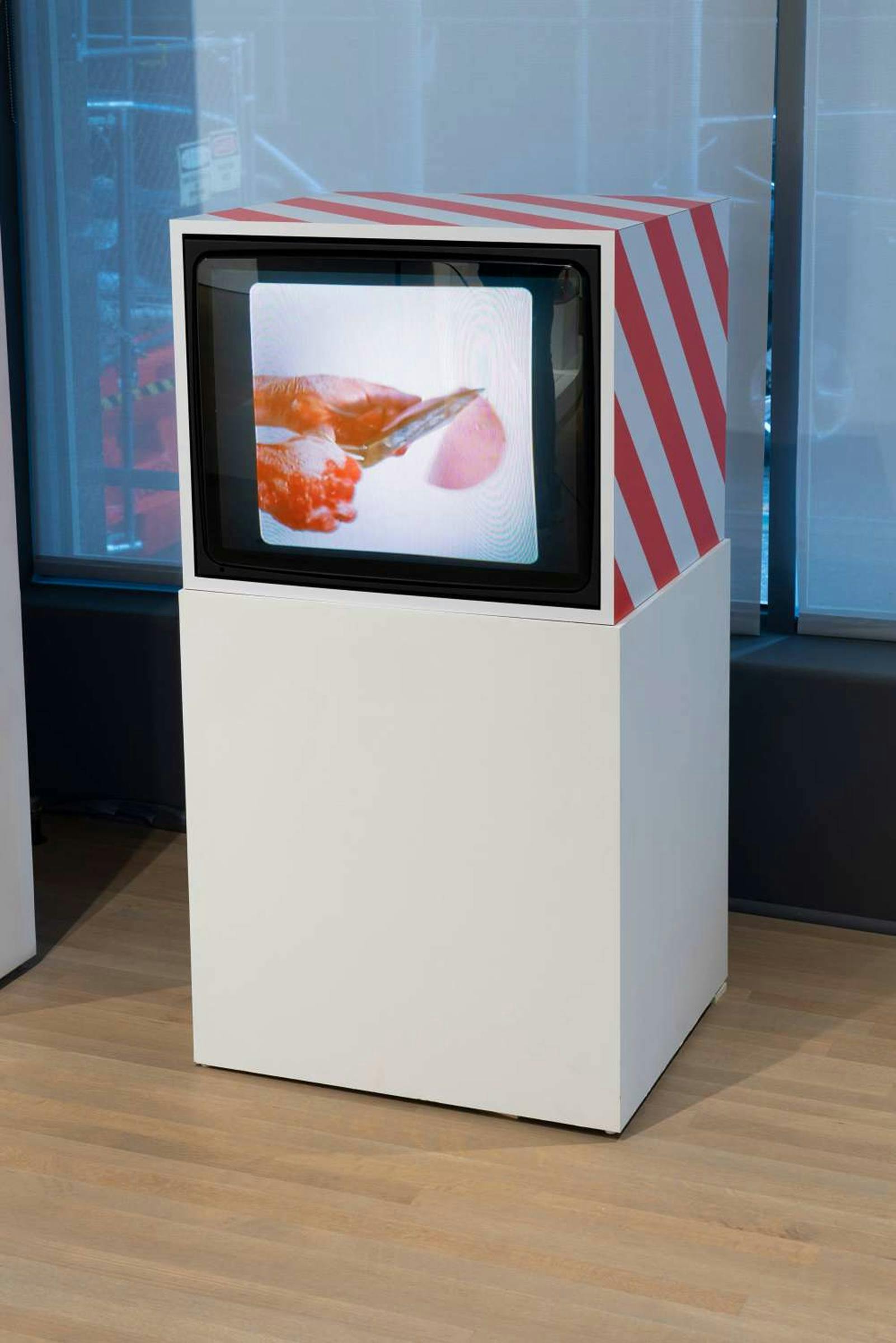 White painted TV stand with red stripes stands in a gallery space. Image of hands cutting through ham on the screen.