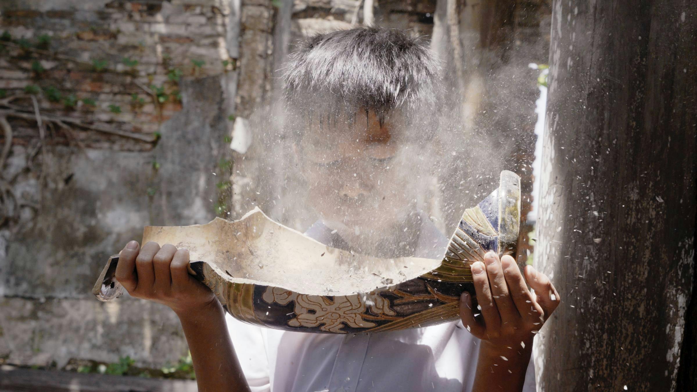 Image of a child holding half a smashed vase in their hand, blowing into it so that dust comes out in a cloud in front of their face. 