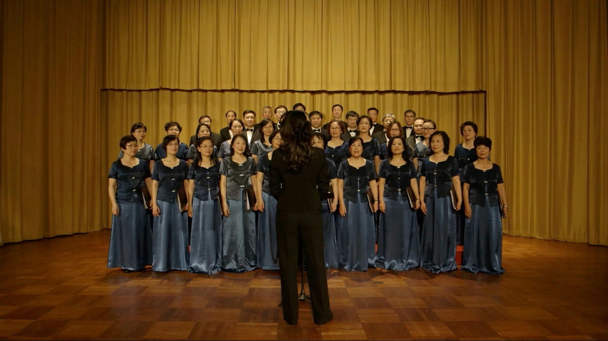 A image of people in Beijing, China singing in a choir. We can see their whole bodies and they are wearing grey floor length dresses