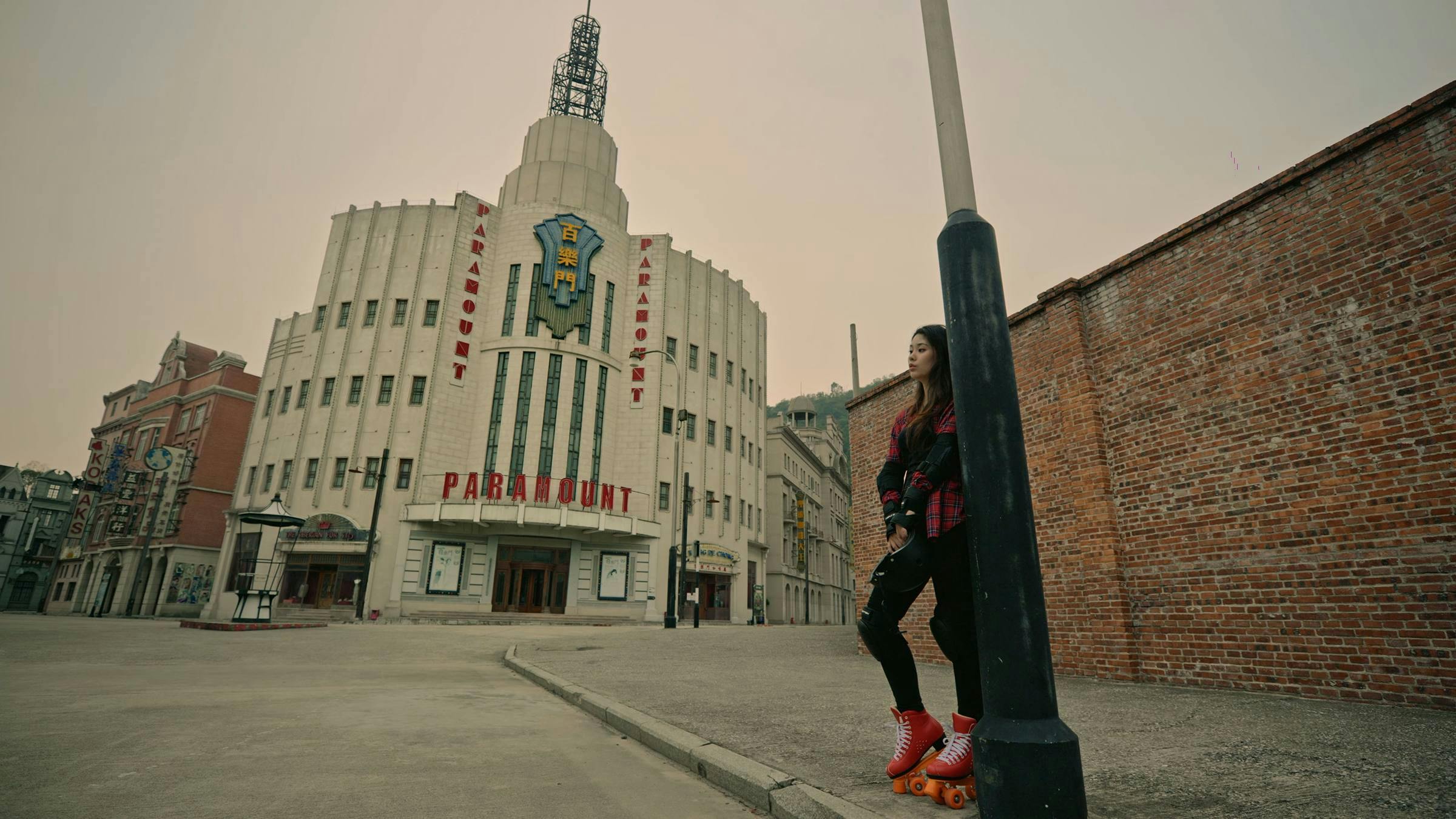 Image of someone wearing orange roller skates stood next to a street lamp on a street corner. A large grey building is in the background and the street is empty.