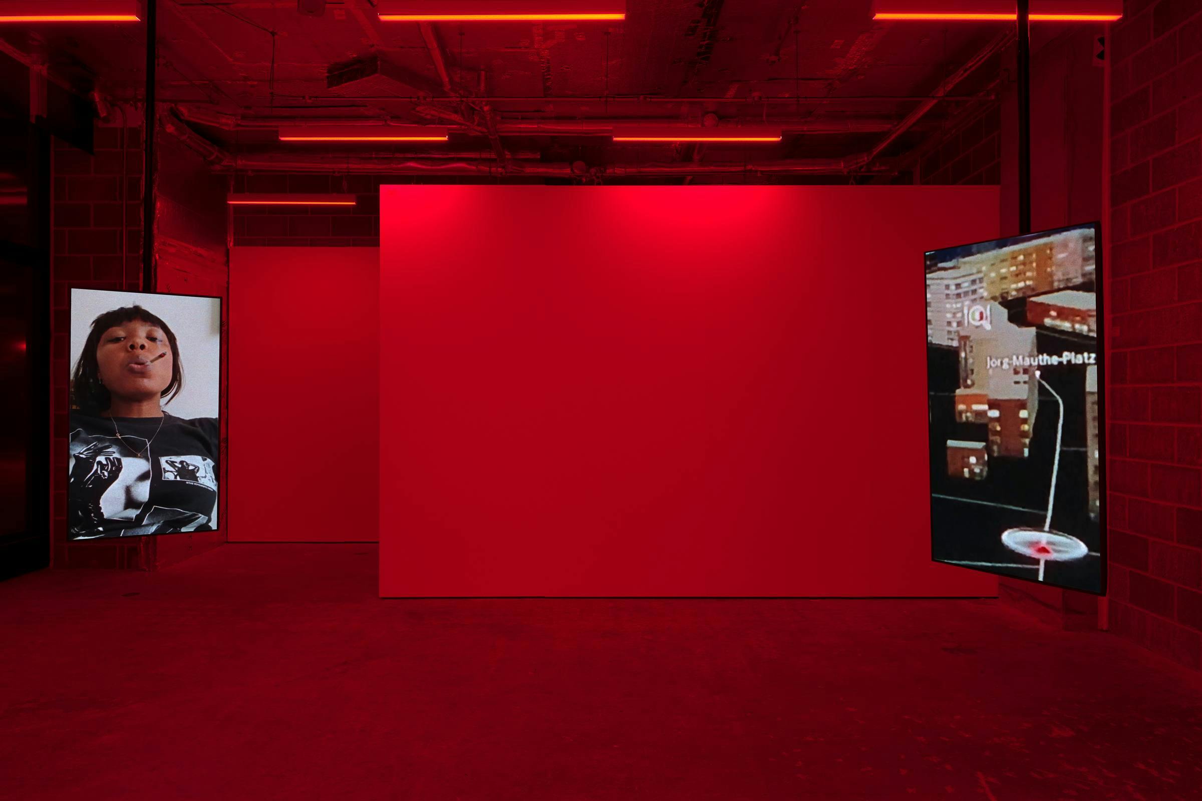 A image of art works in a gallery space. The space is lit red and there are 2 vertical monitors on view that are suspended from the ceiling