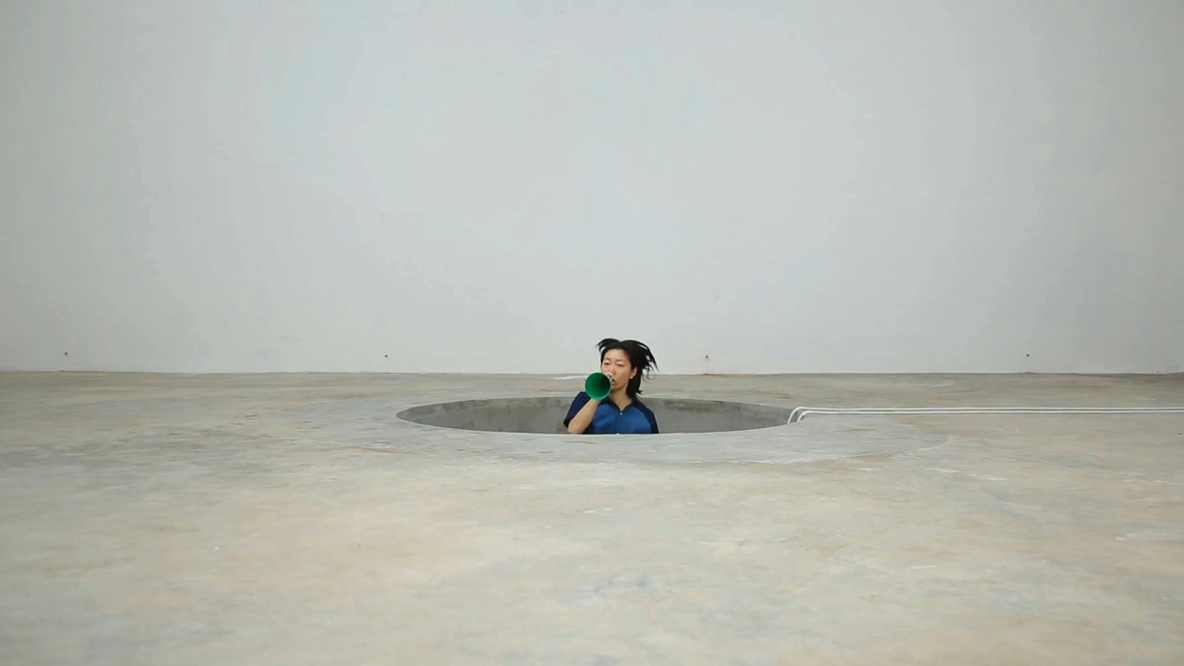 A person blows into a blue horn whilst sticking out of a hole in a concrete floor. The room is painted grey.