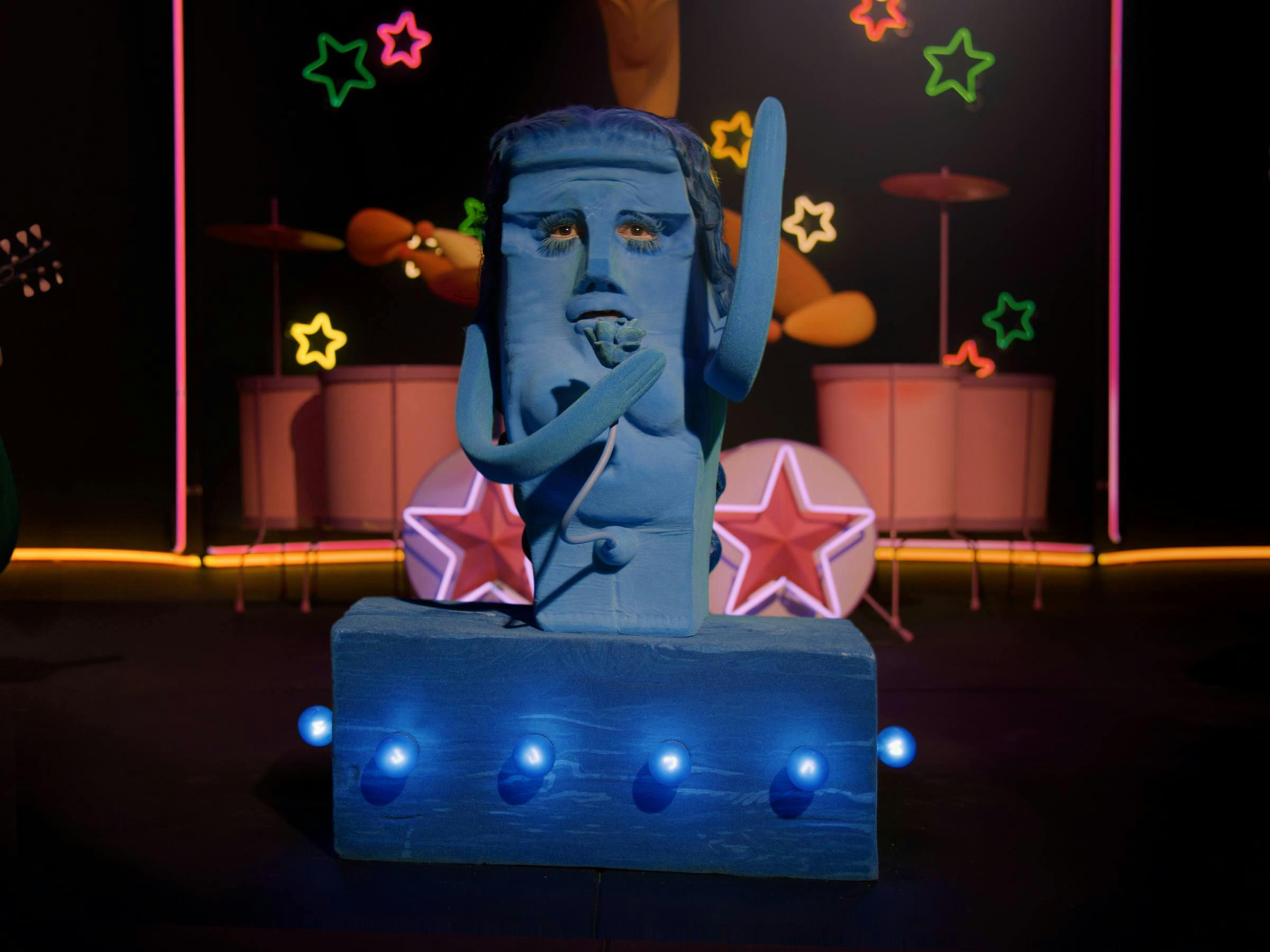 a blue plasticine figure is atop a blue box that has blue light bulbs protruding from it. Pink, yellow and green neon stars are visible as well as 2 drum kits in background. 