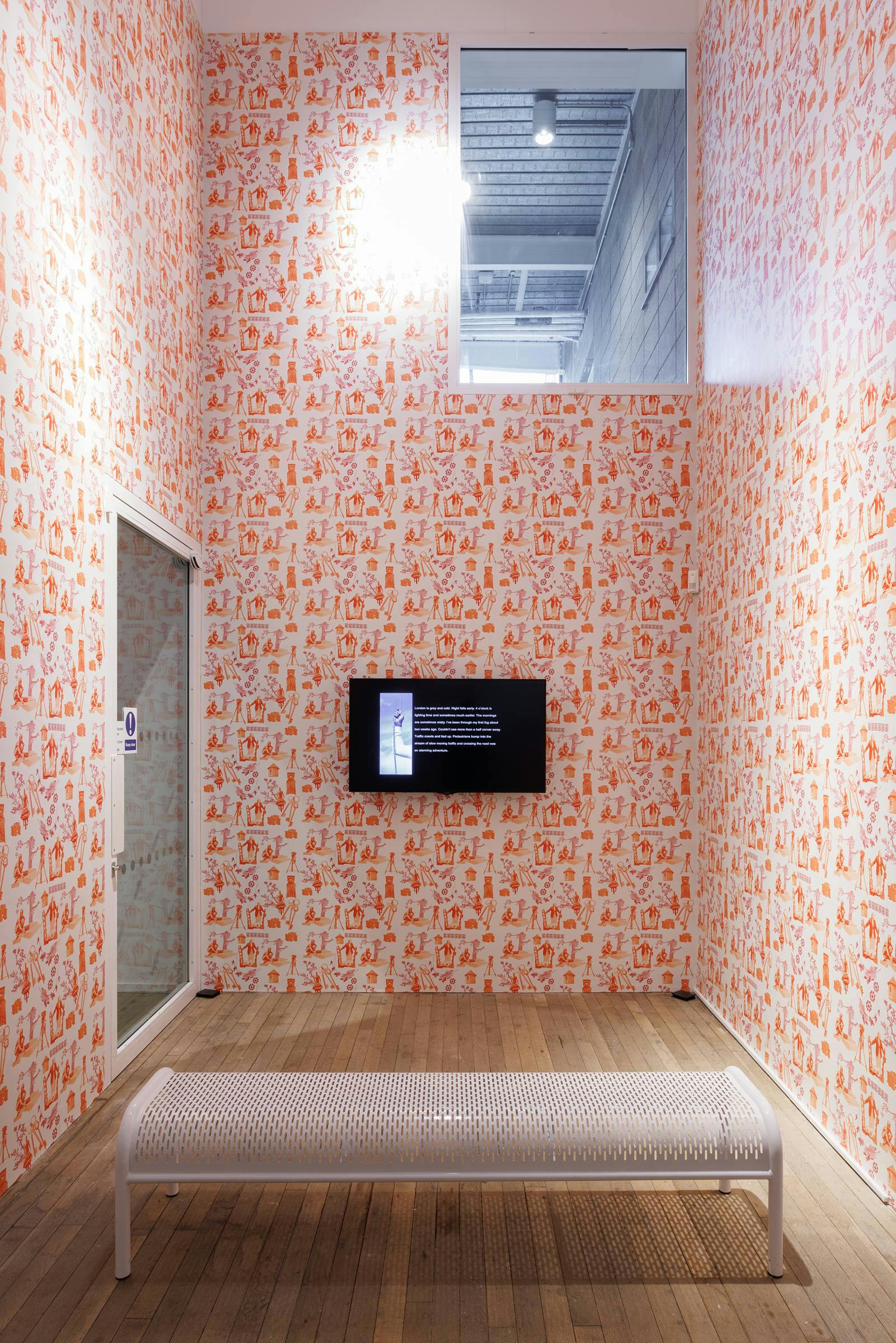 Gallery space with walls covered in orange patterned wallpaper. There is a glass door on the left, a white bench in the centre and a TV mounted on the far wall.